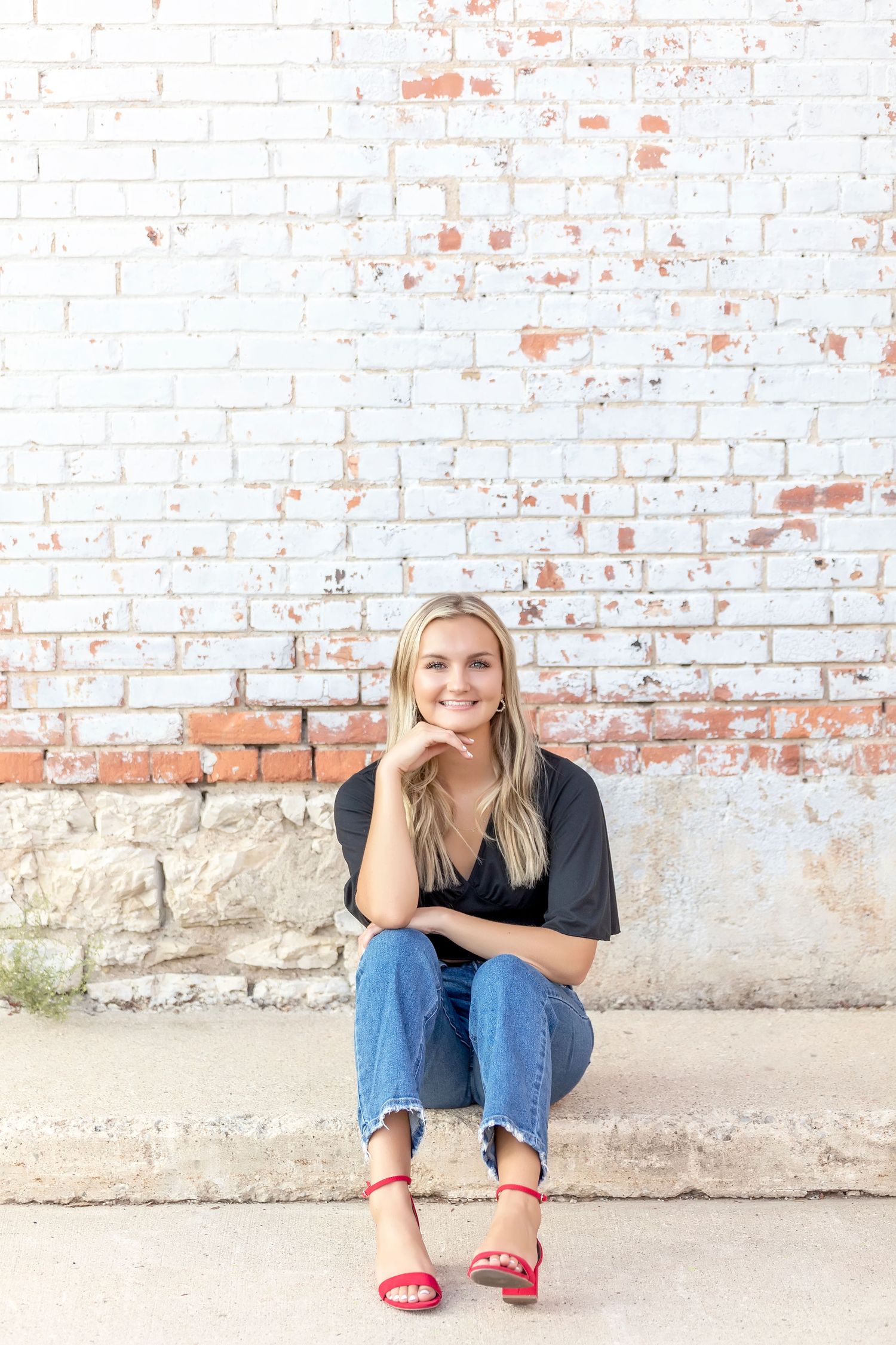 Senior portrait of girl seated against white brick wall in Mason City wearing red sandals and jeans.
