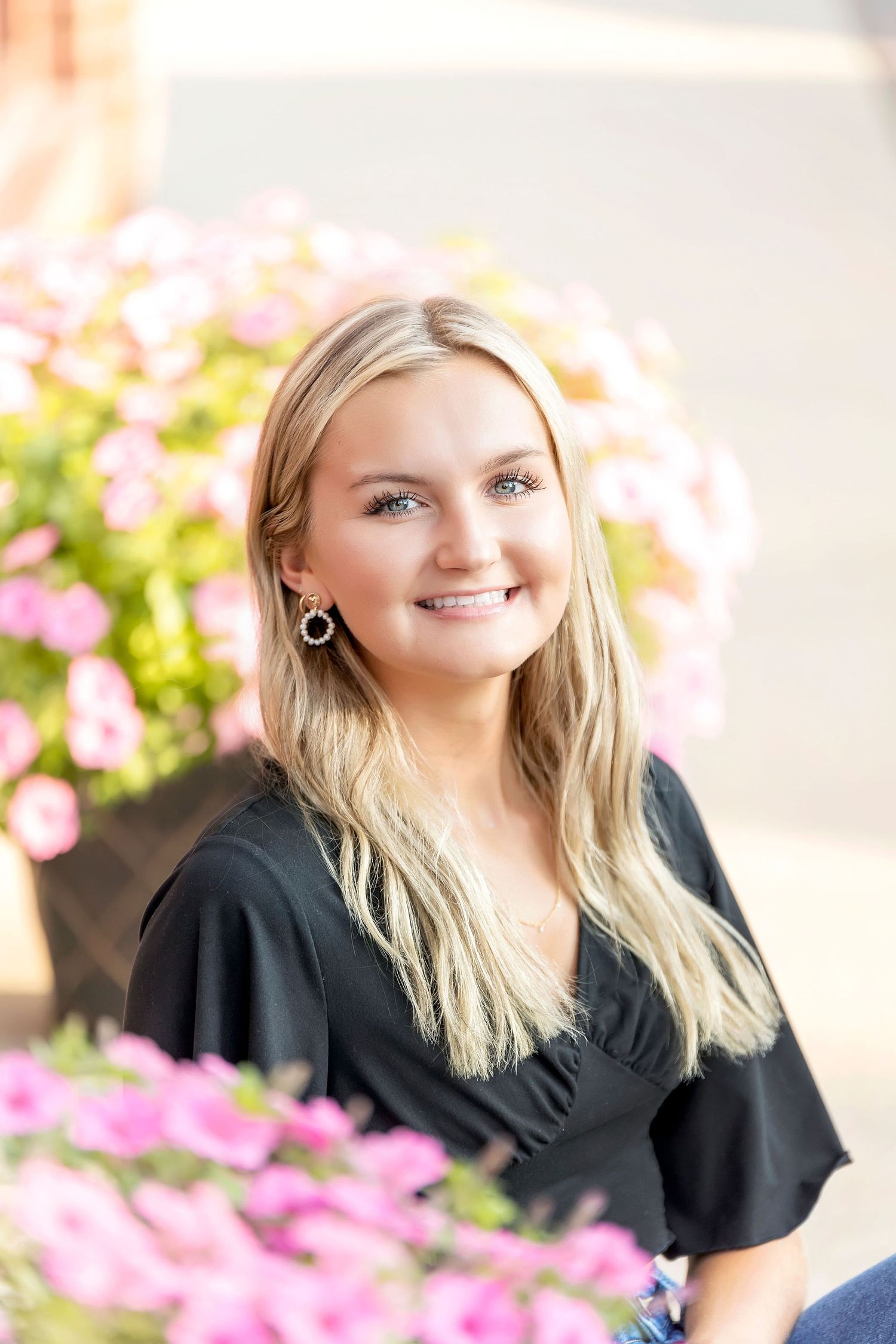 Portrait photo with pink flowers in foreground showing someone in a black top with long blonde hair and silver earrings.