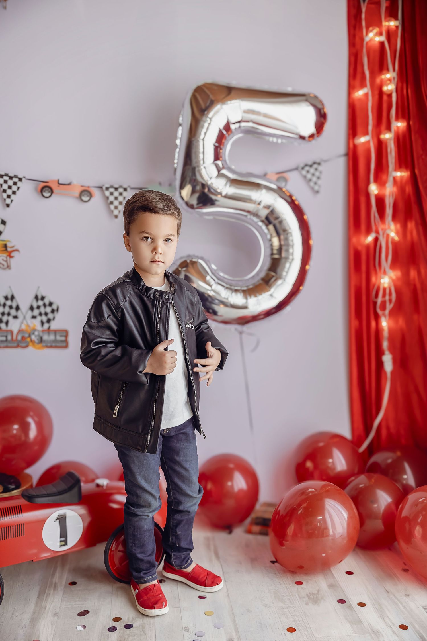 Un nino guapo de 5 anos posa para la camara con una chaqueta negra de cuero hay globos rojos en el piso y un globo en forma del numero 5