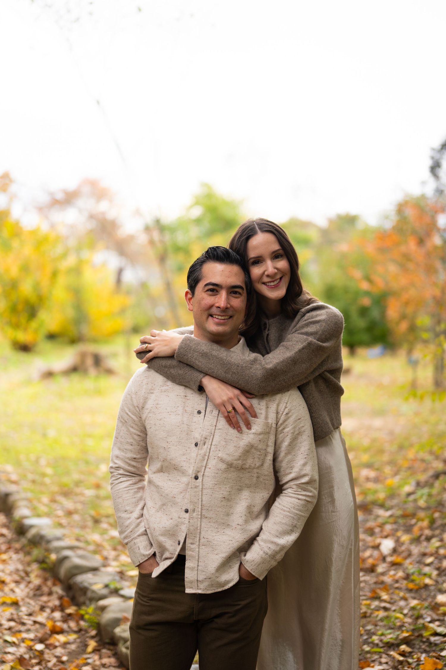 Smiling couple embracing in an autumn park, woman hugging man from behind among yellow fall foliage.
