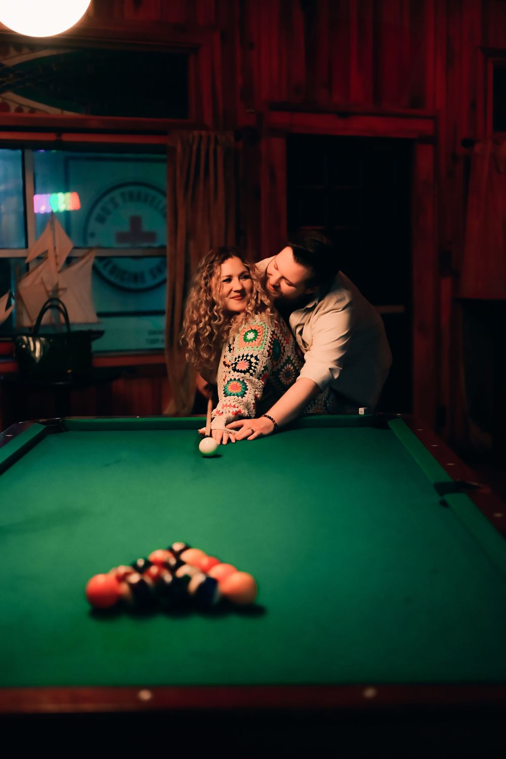 A couple shares an intimate moment while playing pool at a dimly lit bar with red walls and a green felt table.