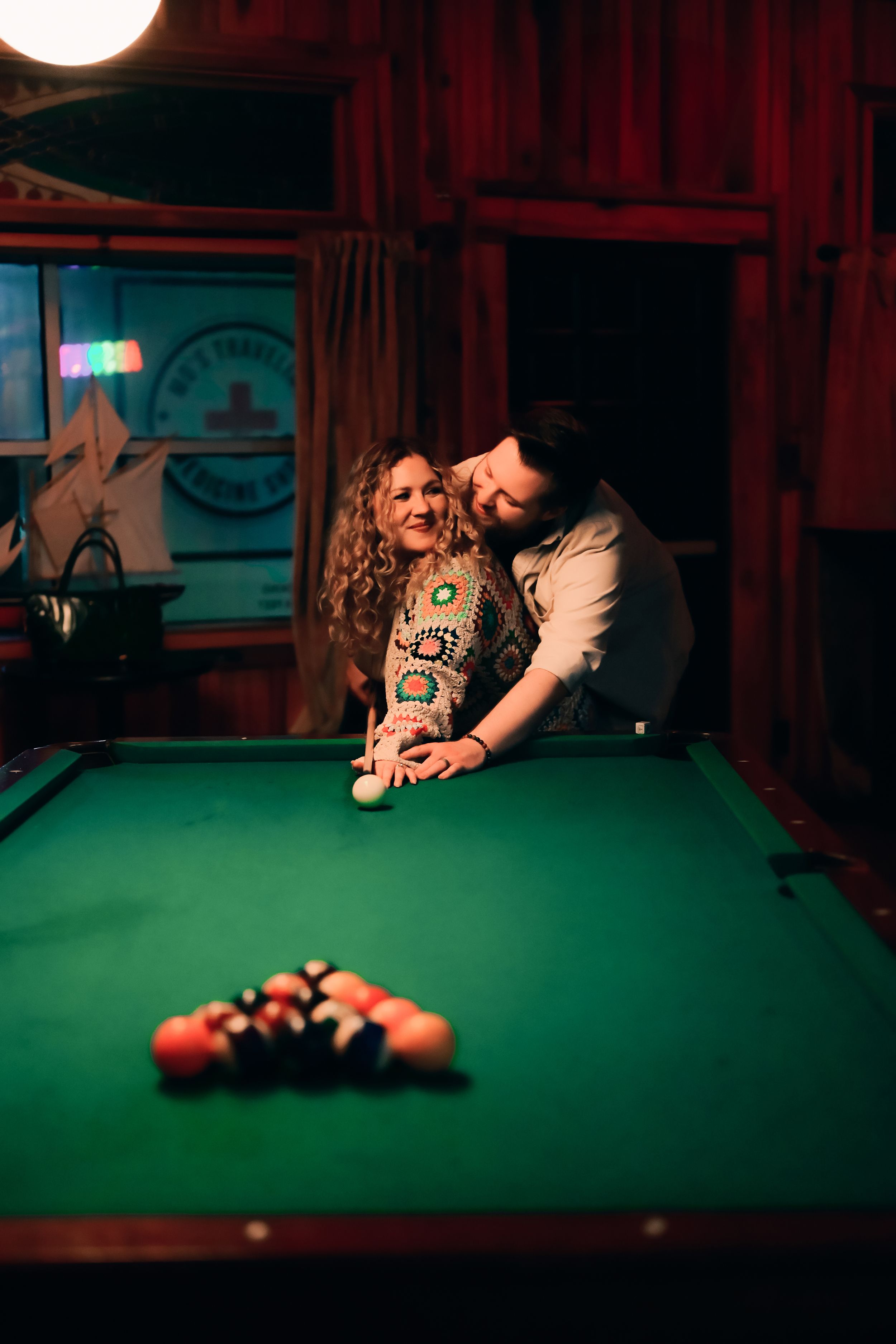 A couple shares an intimate moment while playing pool at a dimly lit bar with red walls and a green felt table.