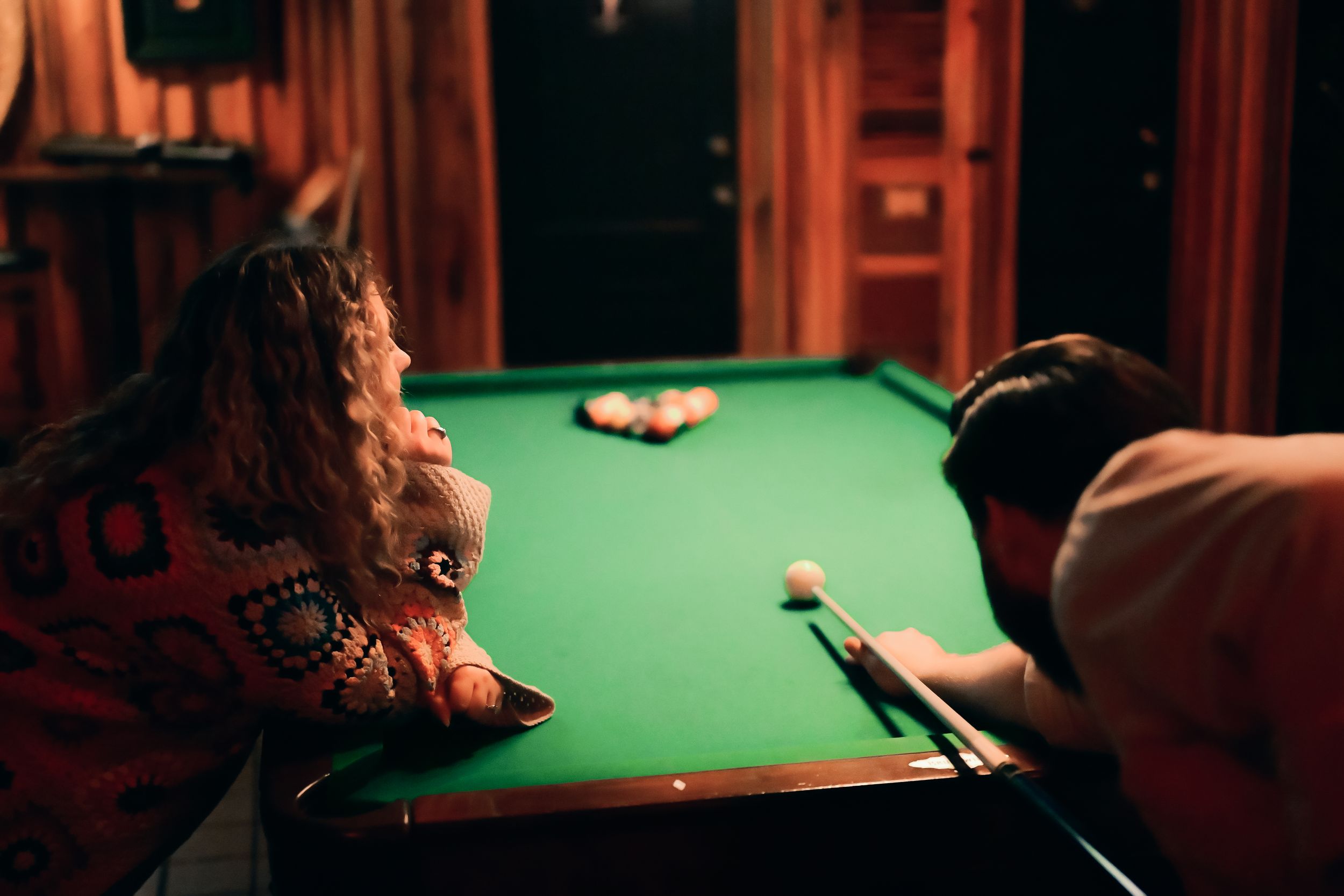 Players lean over a green pool table during a competitive billiards match in a dimly lit room.
