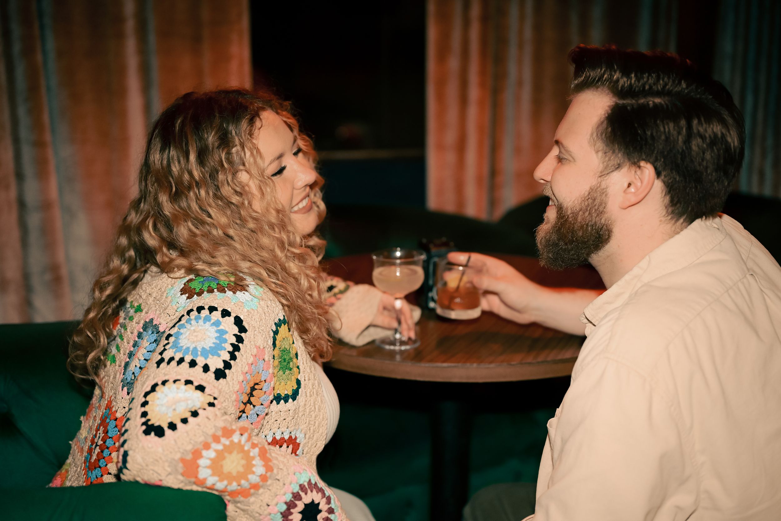 Two people share drinks and laughter while sitting at a dimly lit bar table.