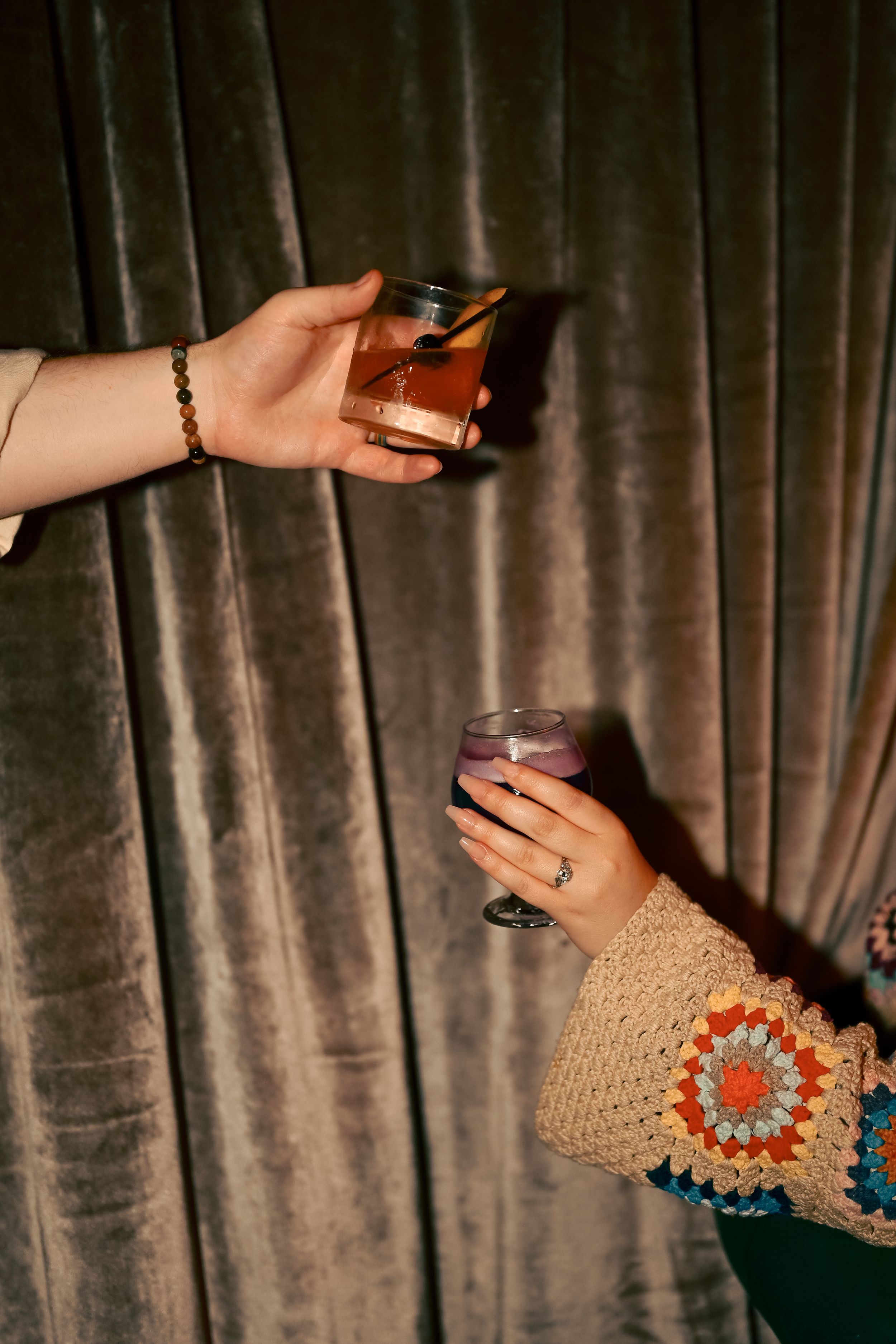 Two hands holding drinks against a gray velvet curtain backdrop, one wearing a cozy knit sweater with orange accents.
