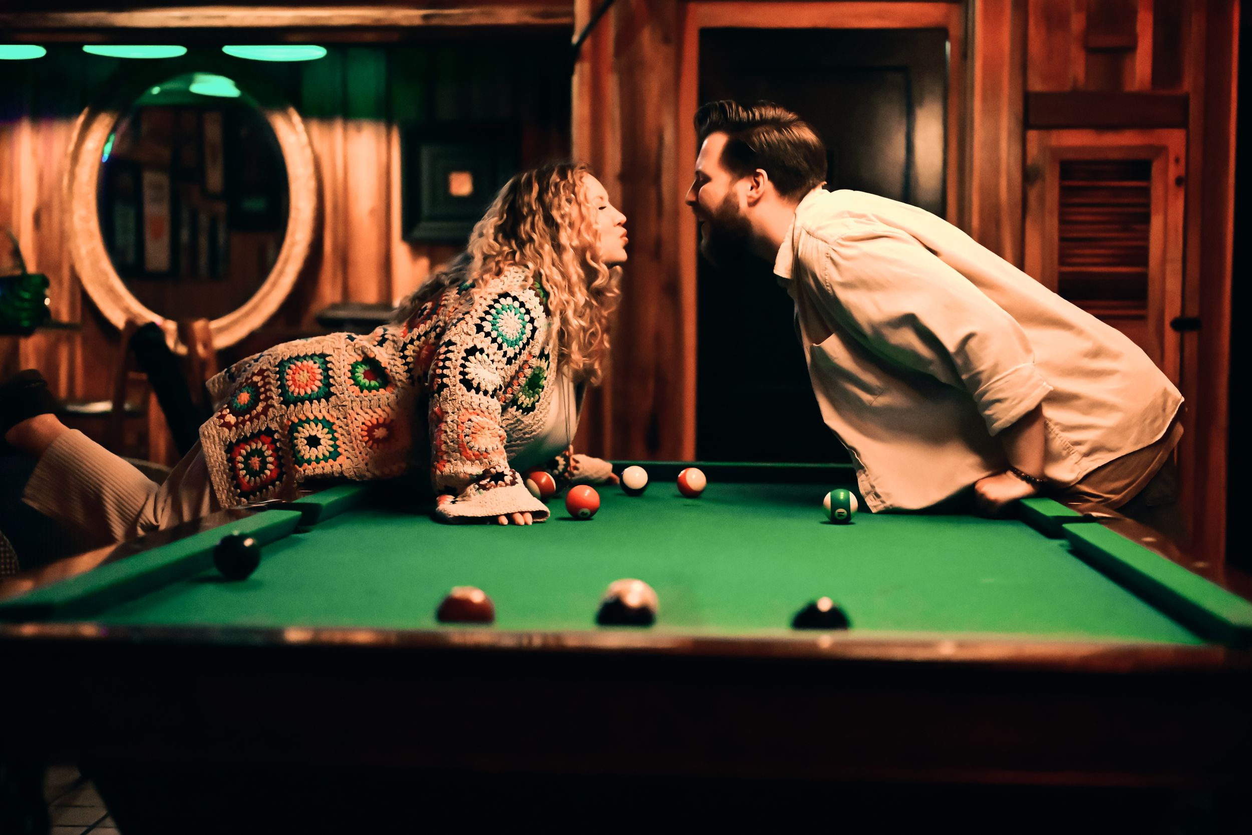 Two people lean across a pool table during a game in a warmly lit room with wood paneling.