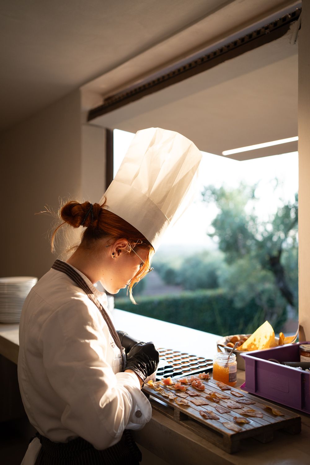 Chef in white uniform and hat carefully prepares food at a commercial kitchen counter in natural lighting.