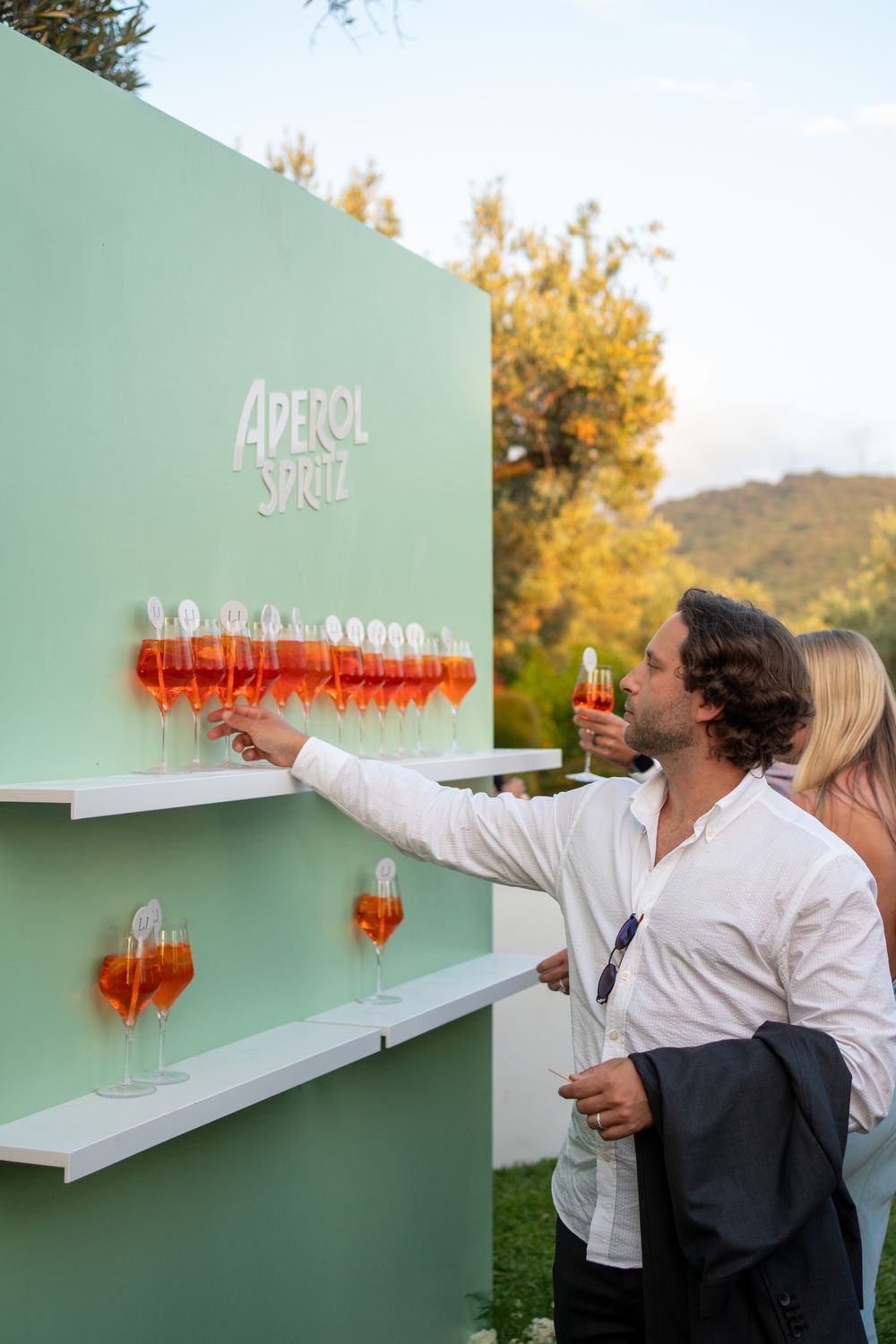 A series of aperol spritz cocktails arranged in a row on a mint green wall-mounted shelf at an outdoor event.