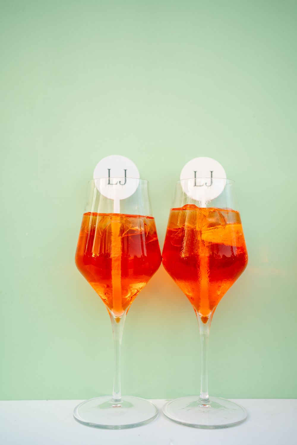Two stemmed wine glasses filled with bright orange Aperol Spritz cocktails against a mint green background.