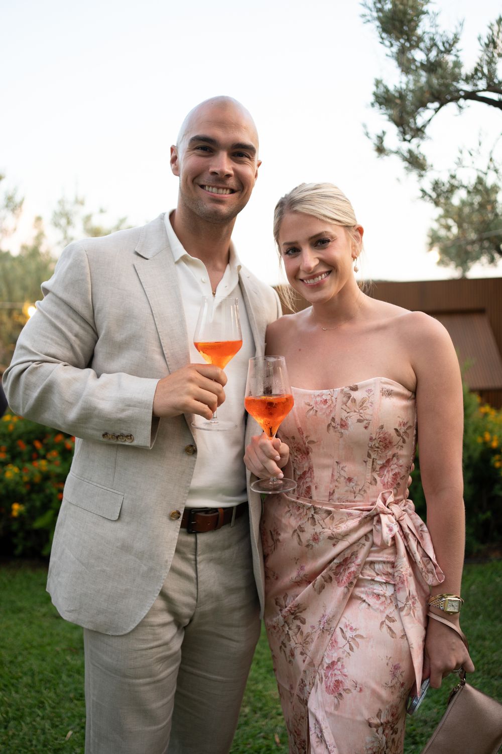 A couple in formal attire holding orange cocktails at an outdoor garden party.