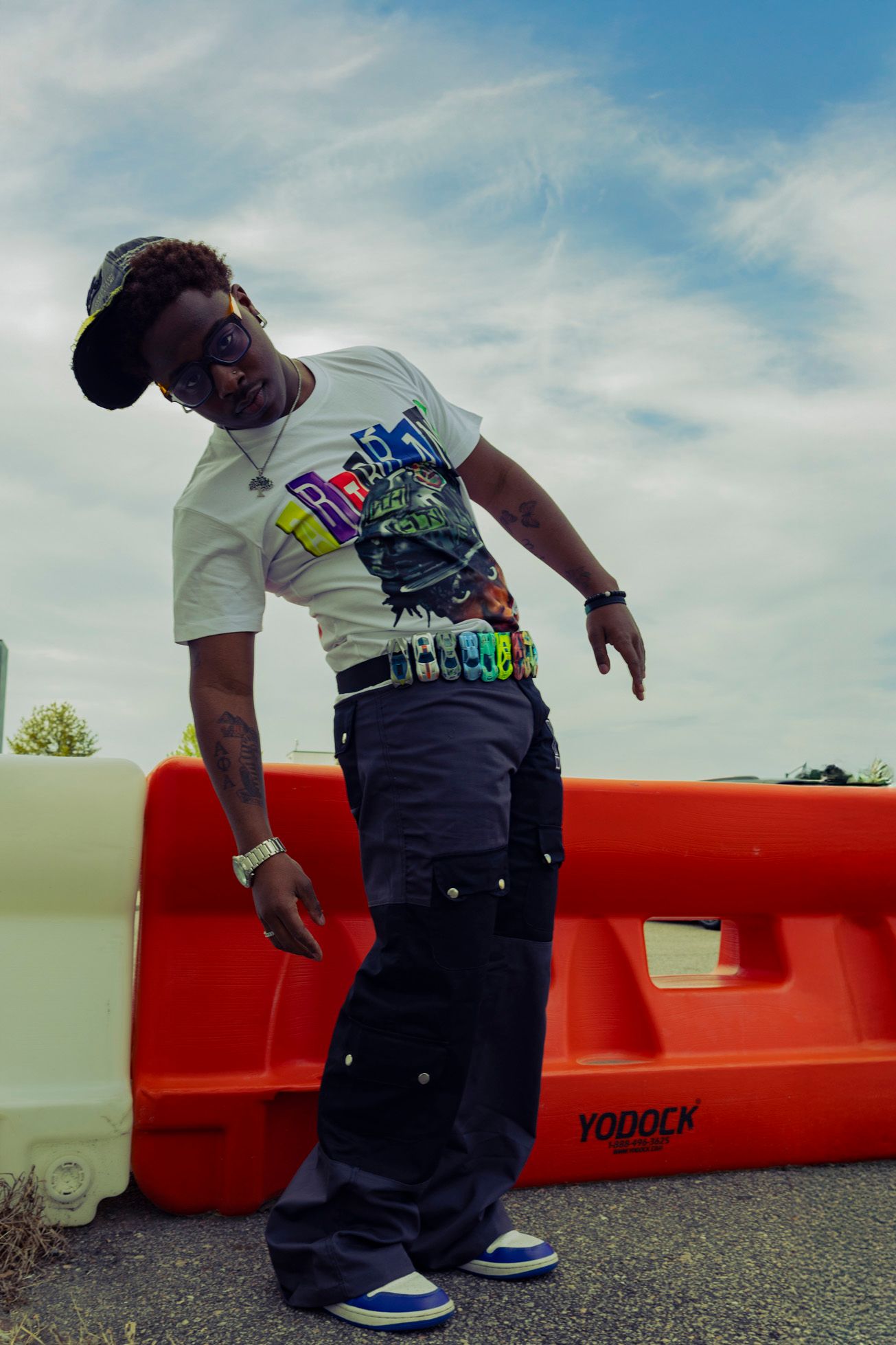 Person in graphic t-shirt and baseball cap posing against orange barrier outdoors.