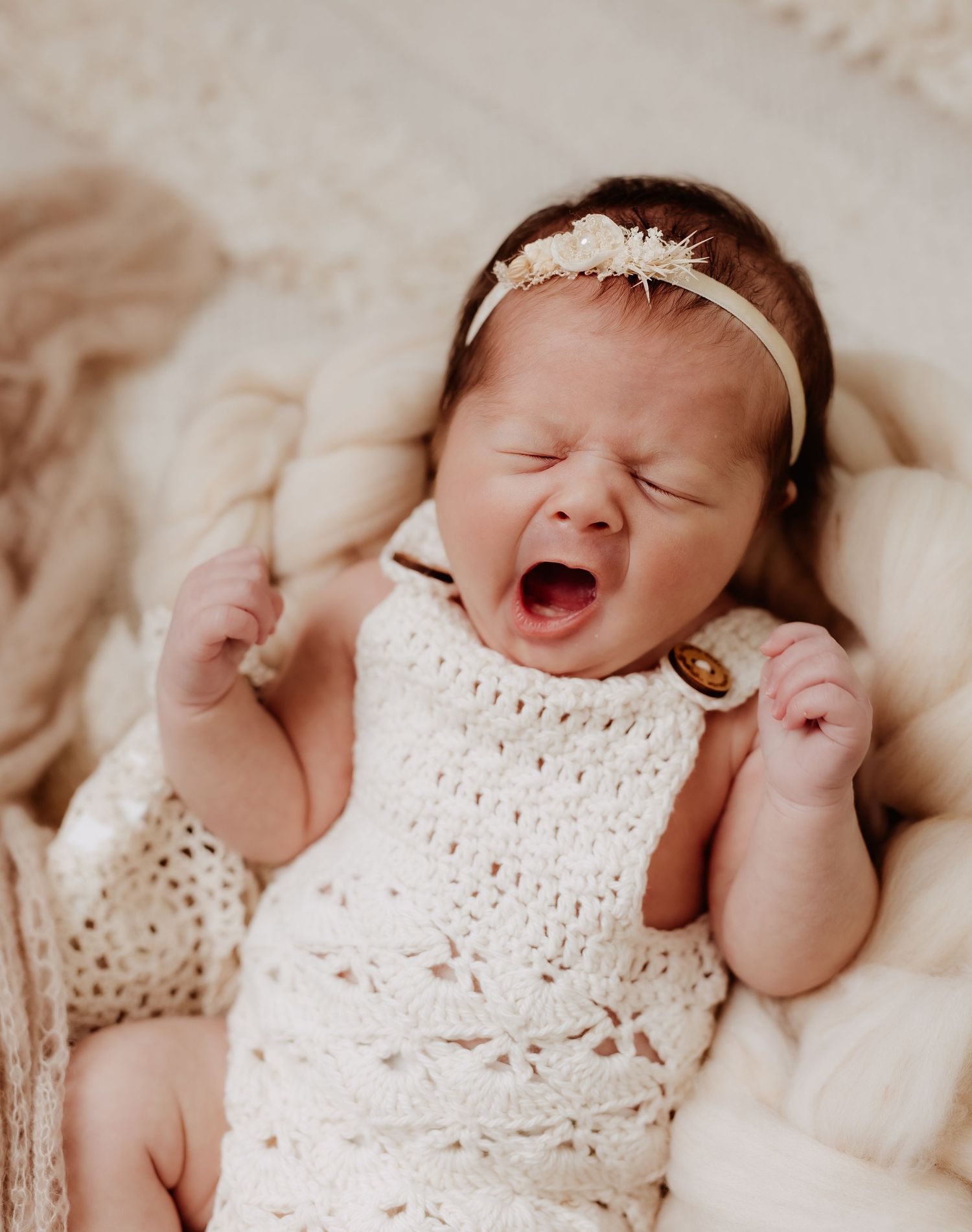 Newborn baby yawning while wearing white lace outfit and headband.