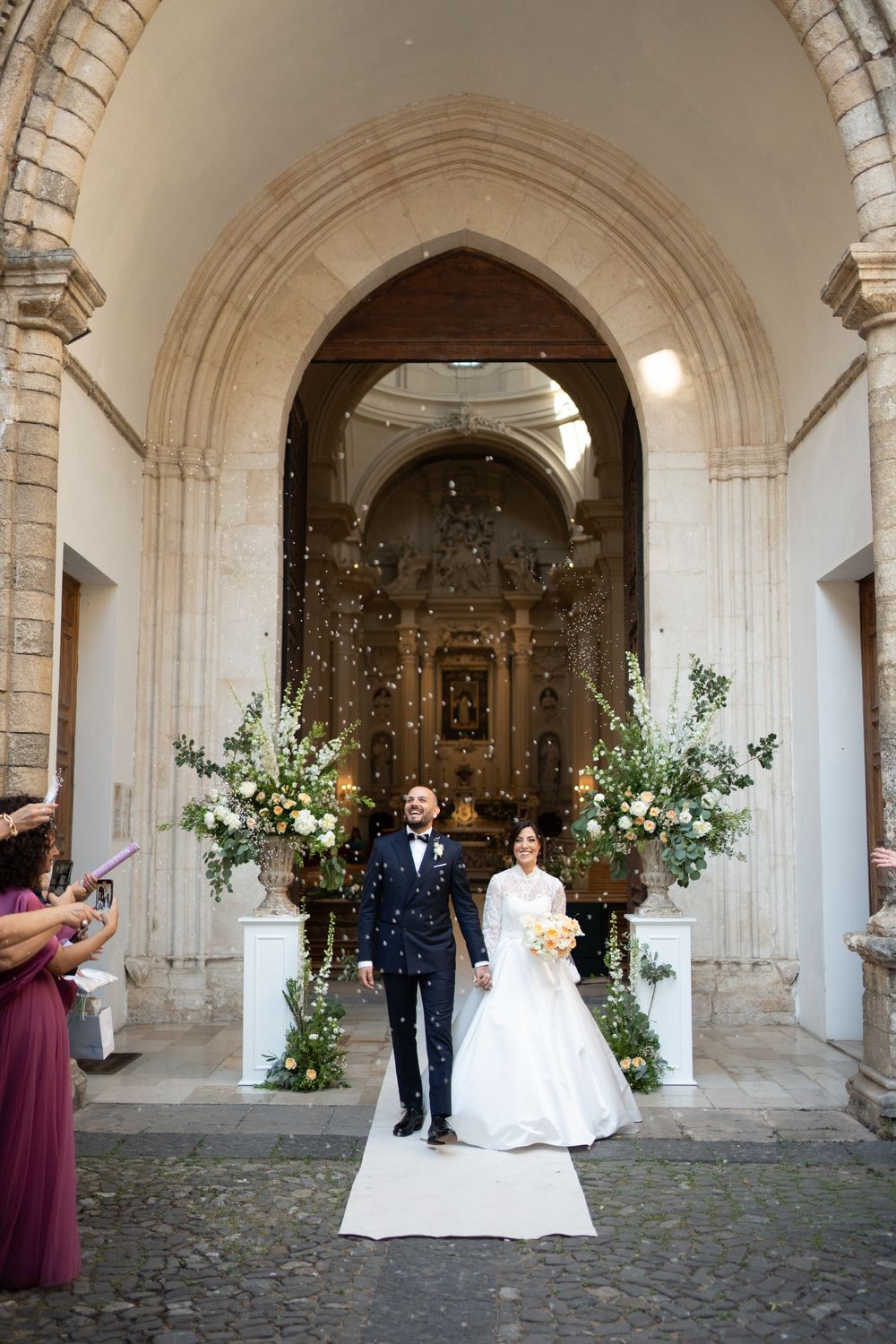 Wedding couple poses under stone archways decorated with floral arrangements at an elegant historic venue.