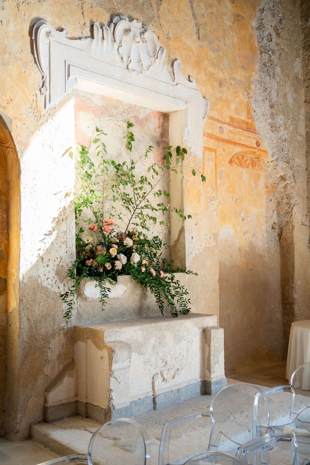 Elegant floral arrangement in a rustic stone alcove with ornate white trim and peach-colored walls in an old Italian villa.