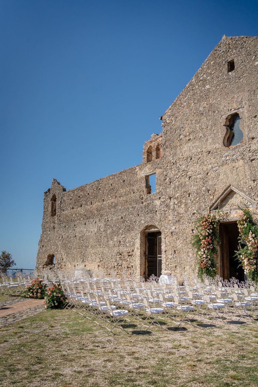 White chairs arranged in rows for an outdoor ceremony against a rustic stone wall of an ancient castle or fortress.