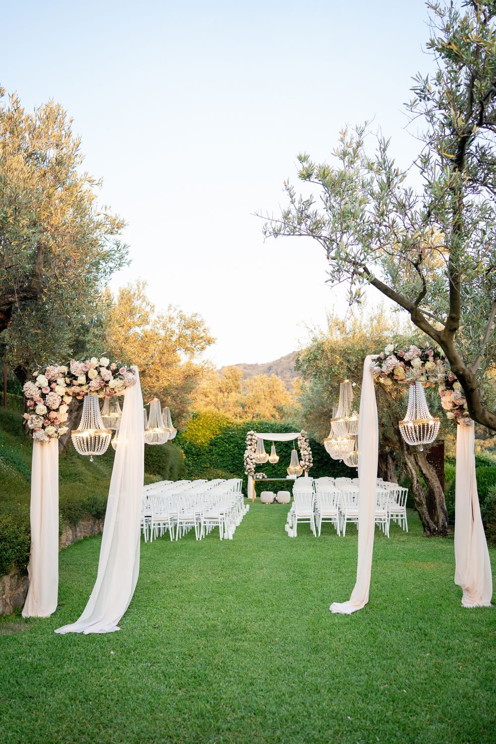 Elegant outdoor wedding ceremony setup with white chairs and floral arrangements on pillars along a grassy aisle.