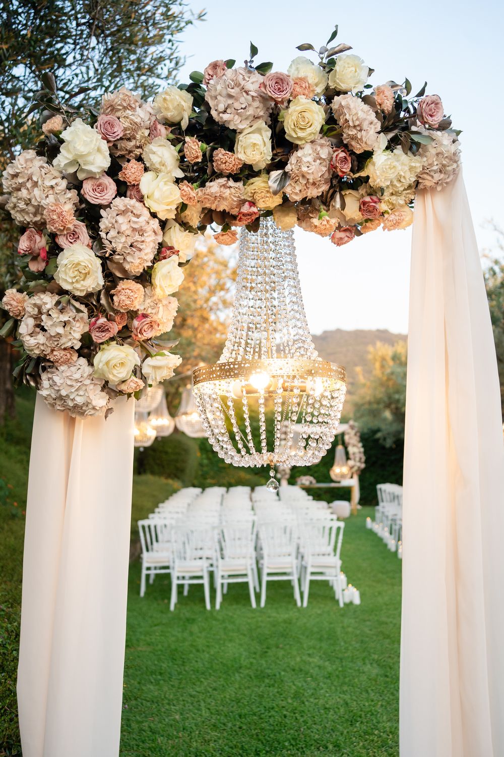 Elegant outdoor wedding ceremony setup with crystal chandelier and floral arch decorated in blush and cream roses.