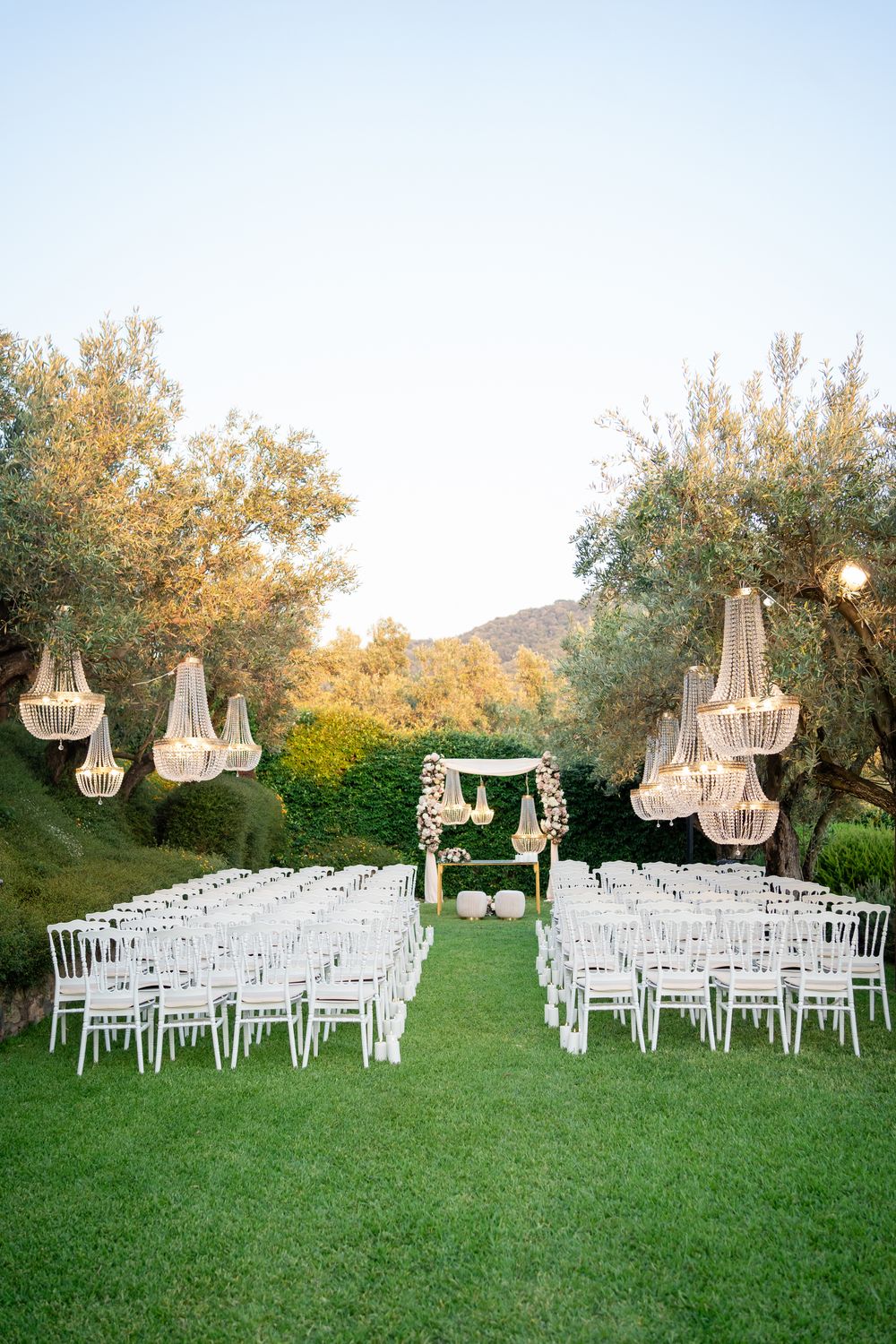 Elegant outdoor wedding ceremony setup with white chairs and crystal chandeliers hanging from trees at sunset.