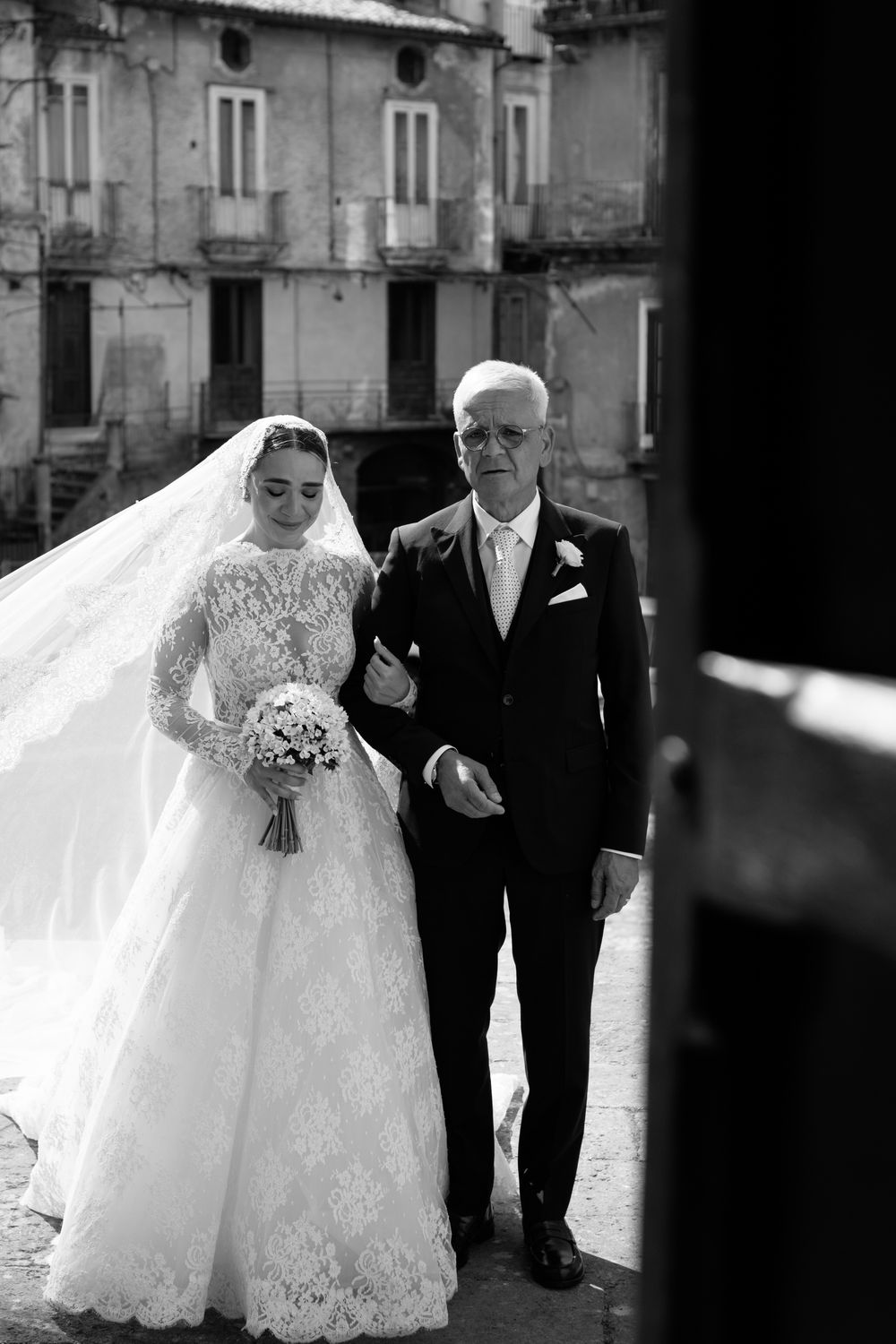 A bride in a white lace wedding dress walks with an escort down a stone street in a traditional ceremony.