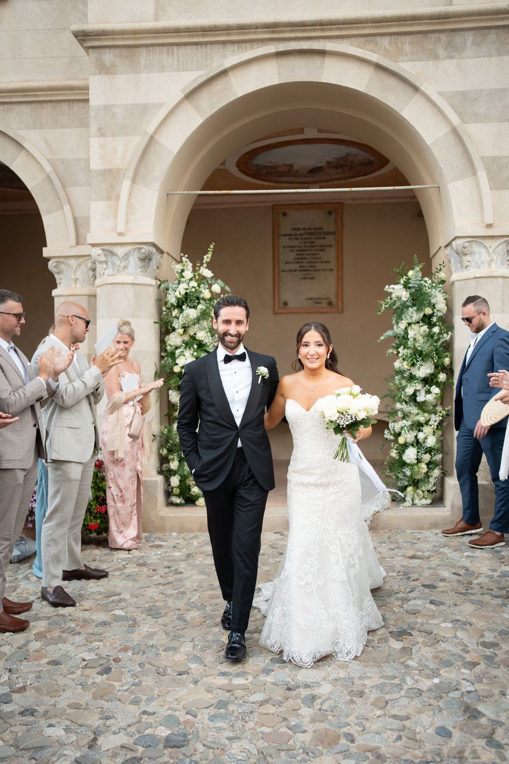 Elegant wedding couple exits stone archway decorated with white floral arrangements as guests look on.