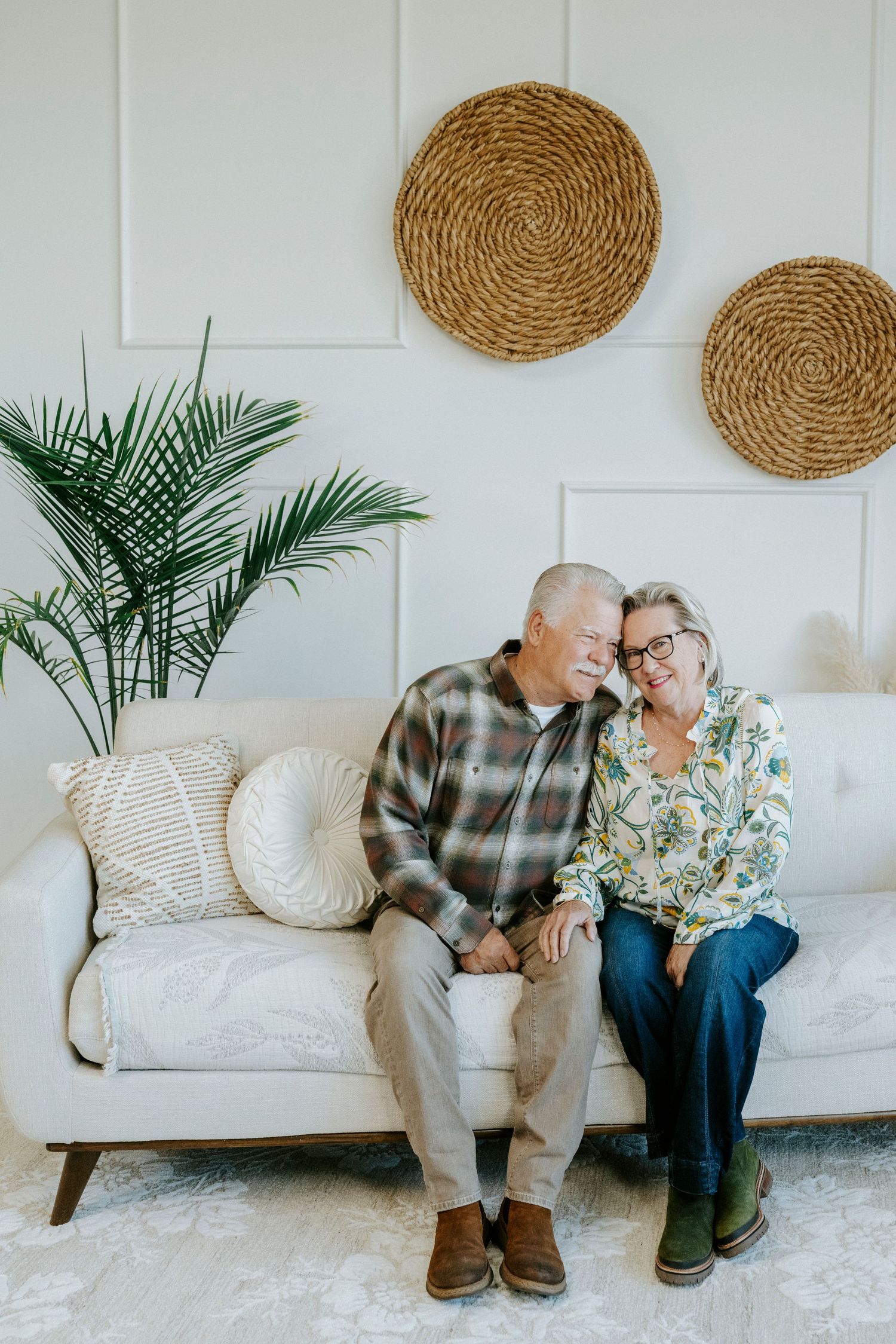 Two people sit together on a white couch with decorative woven baskets and palm fronds on the wall behind them.
