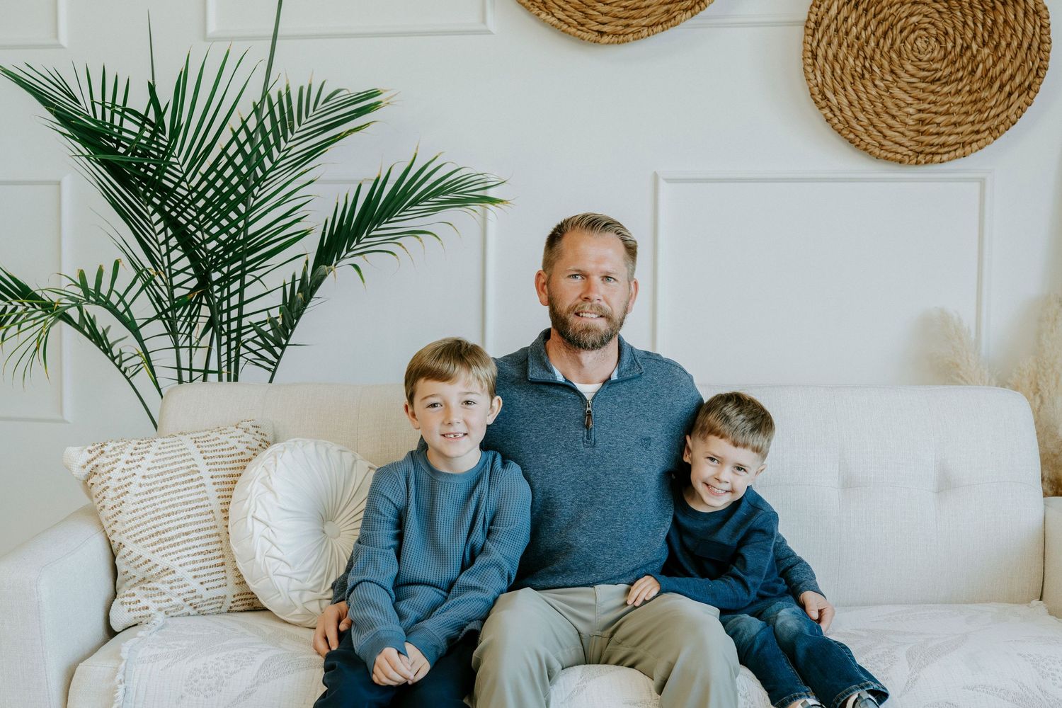 A family wearing coordinated blue and gray outfits poses together on a cream couch with woven wall decor behind them.
