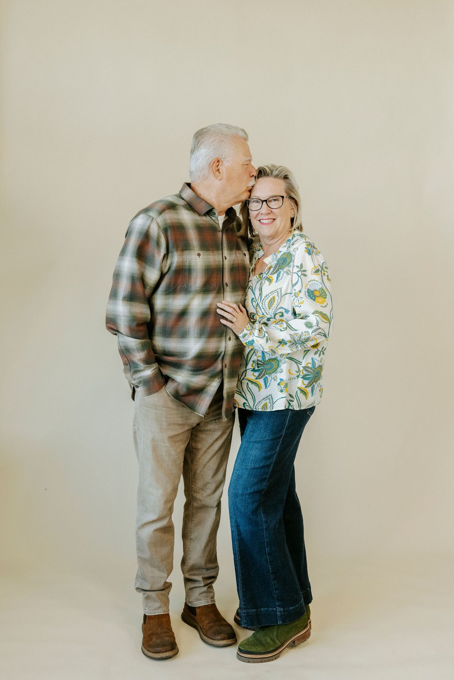 Two elderly people in casual attire share a tender moment against a neutral backdrop.
