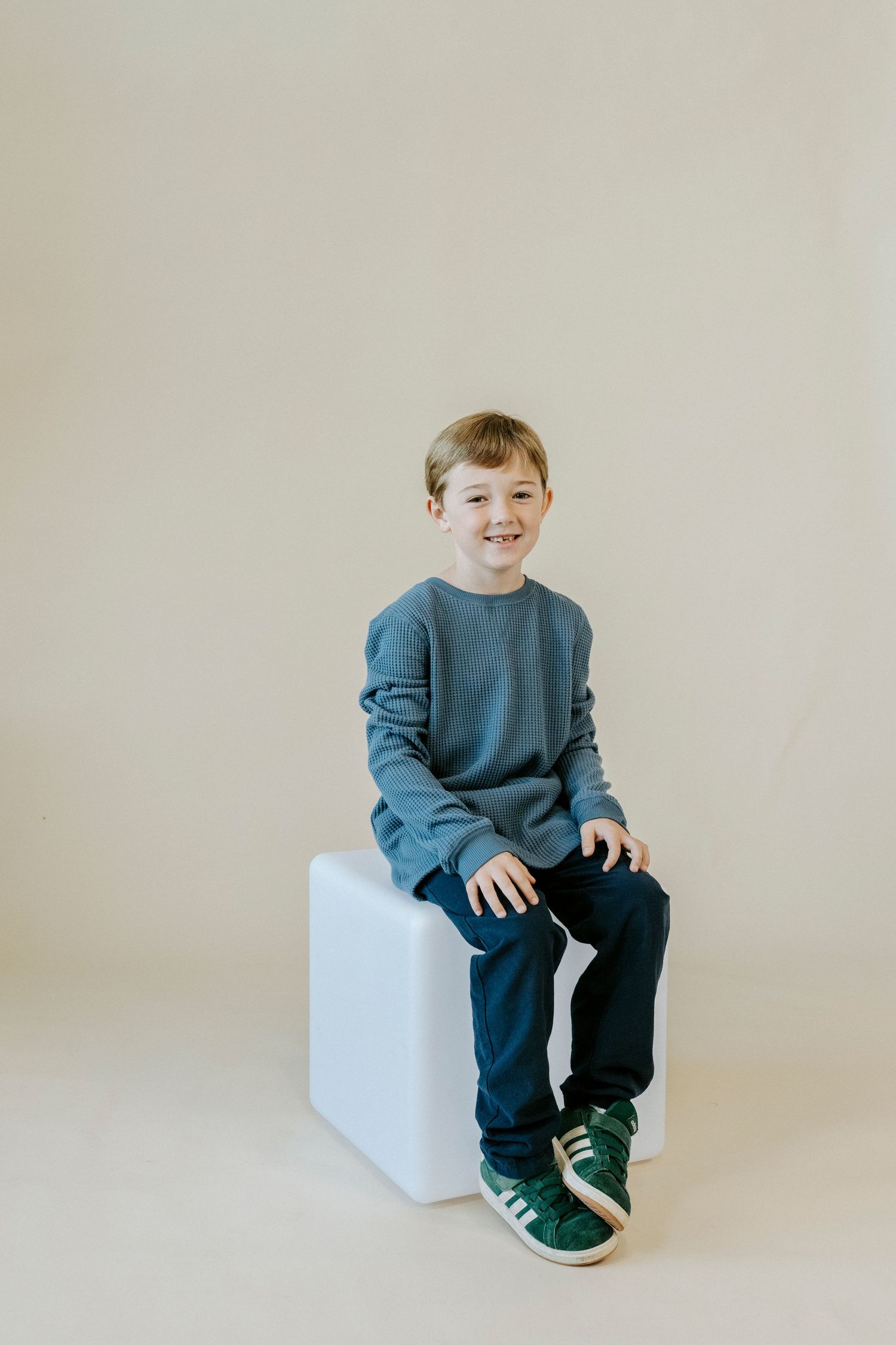 A child in a blue sweater and dark pants sits on a white cube against a neutral background.