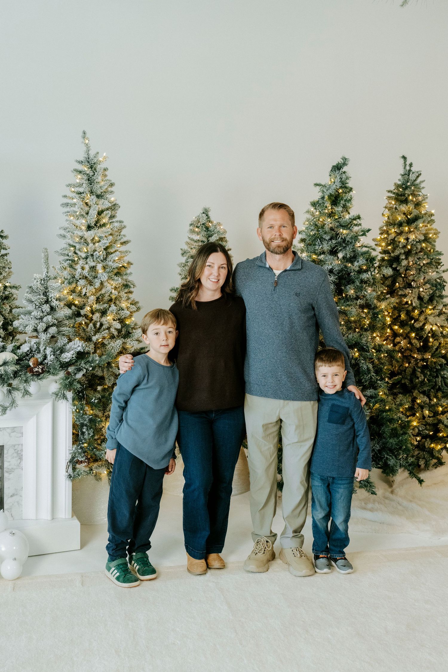 A family poses together for holiday photos in front of lit Christmas trees in a bright white studio setting.