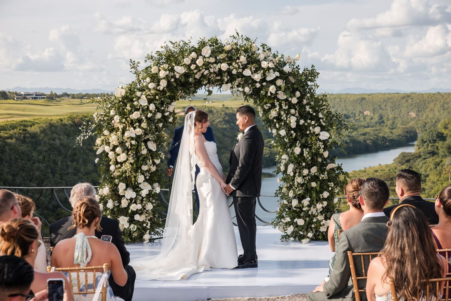 Outdoor wedding ceremony under a beautiful white floral arch overlooking scenic countryside landscape.
