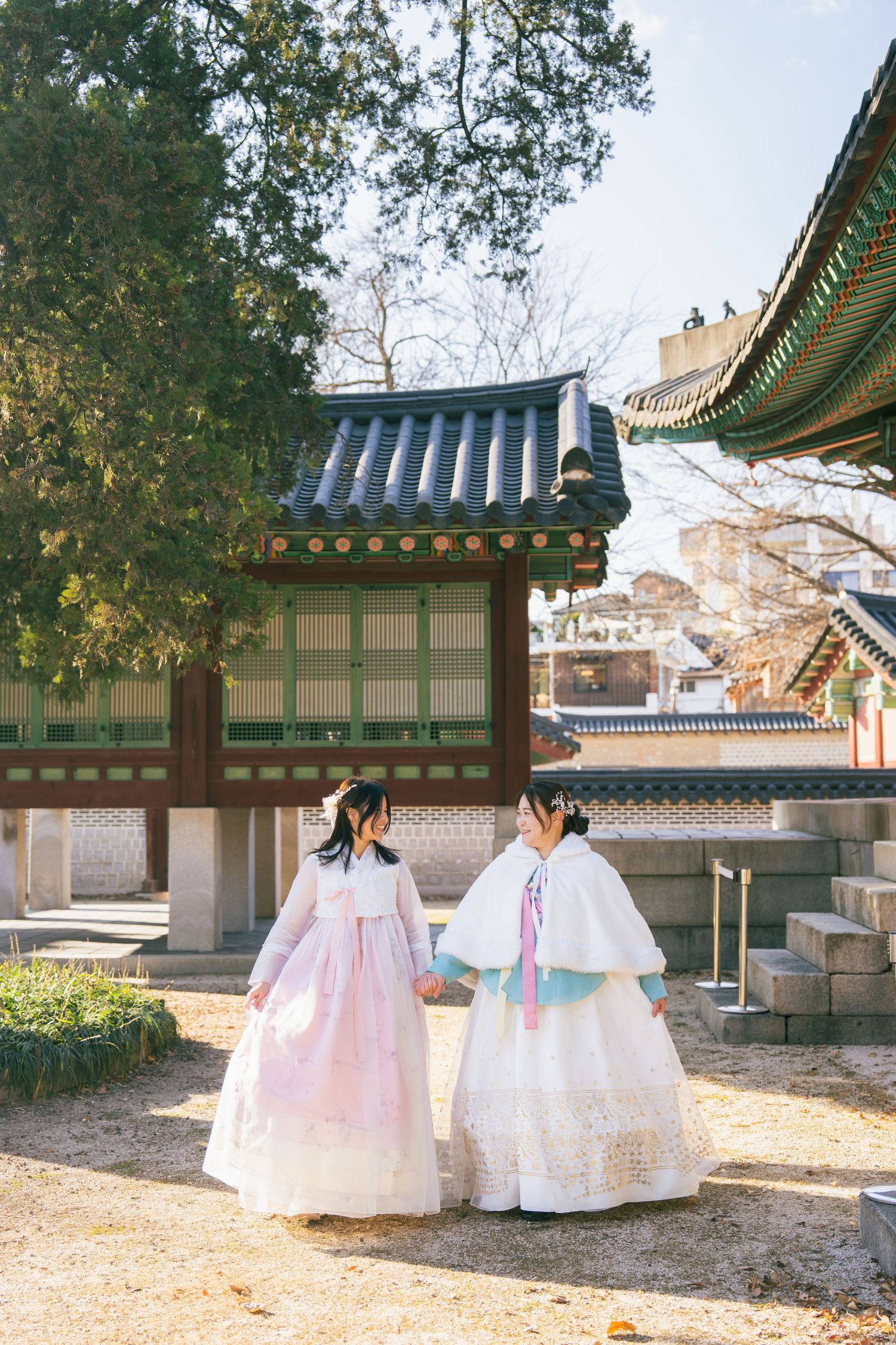 Traditional Korean hanbok dresses shown against historic palace buildings with ornate rooftops in warm sunlight.