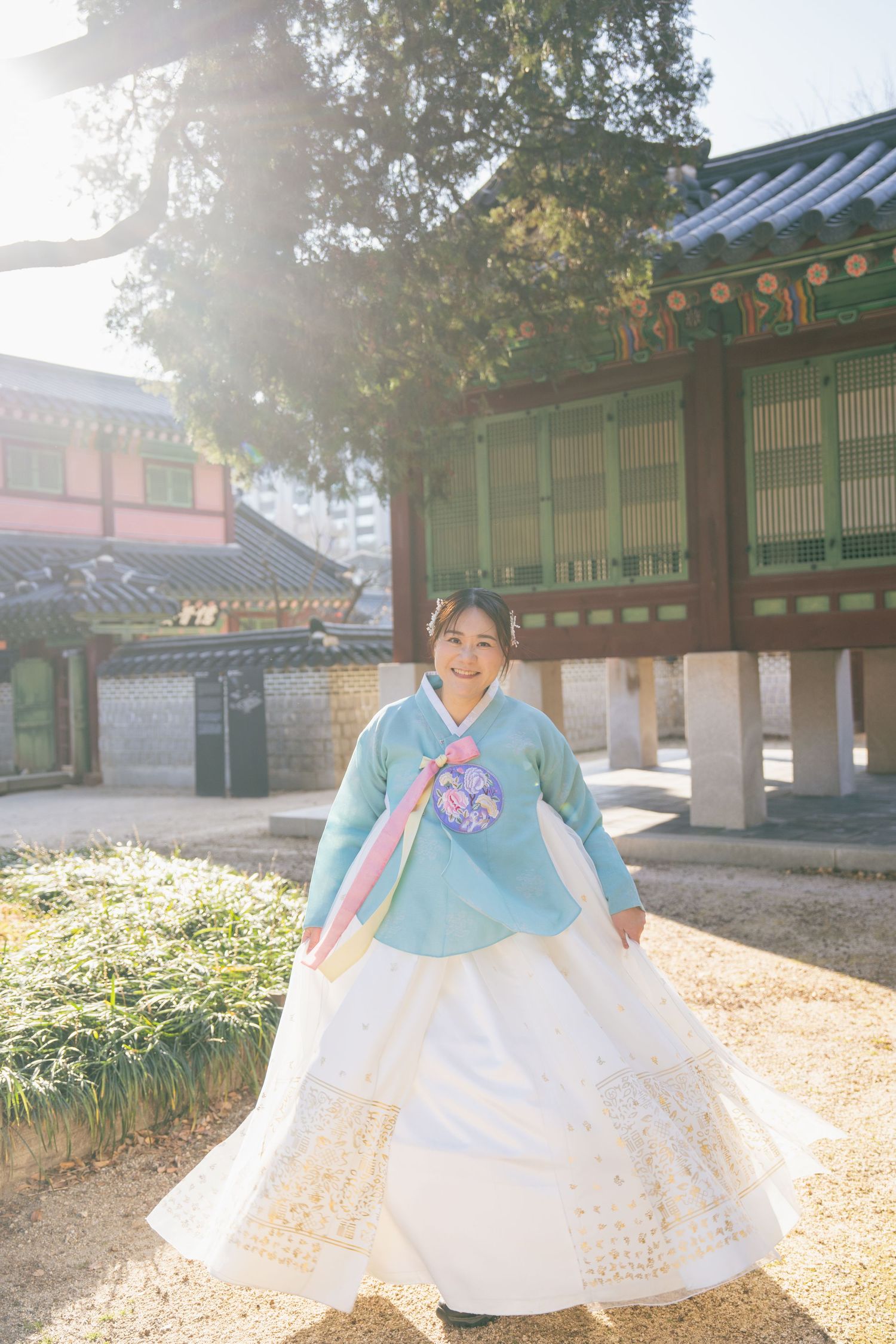 A person wearing a traditional Korean hanbok dress twirls in a sunlit courtyard of a historic temple complex.