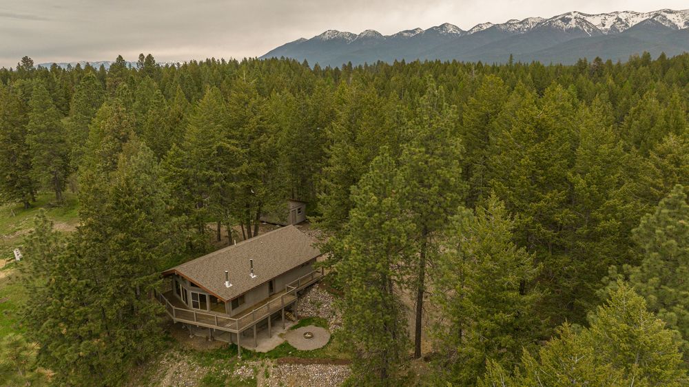 Panoramic mountain landscape with snow-capped peaks, dense forest, and rustic cabin nestled among evergreen trees.