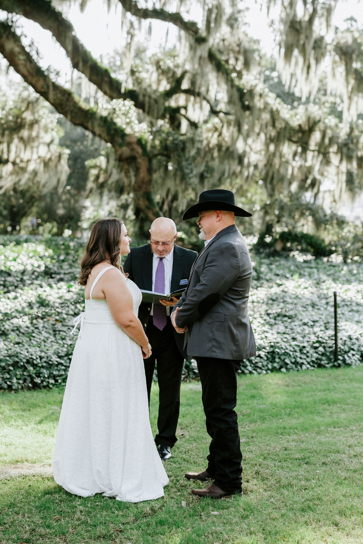 Outdoor wedding ceremony under Spanish moss draped trees with couple and officiant in a garden setting.
