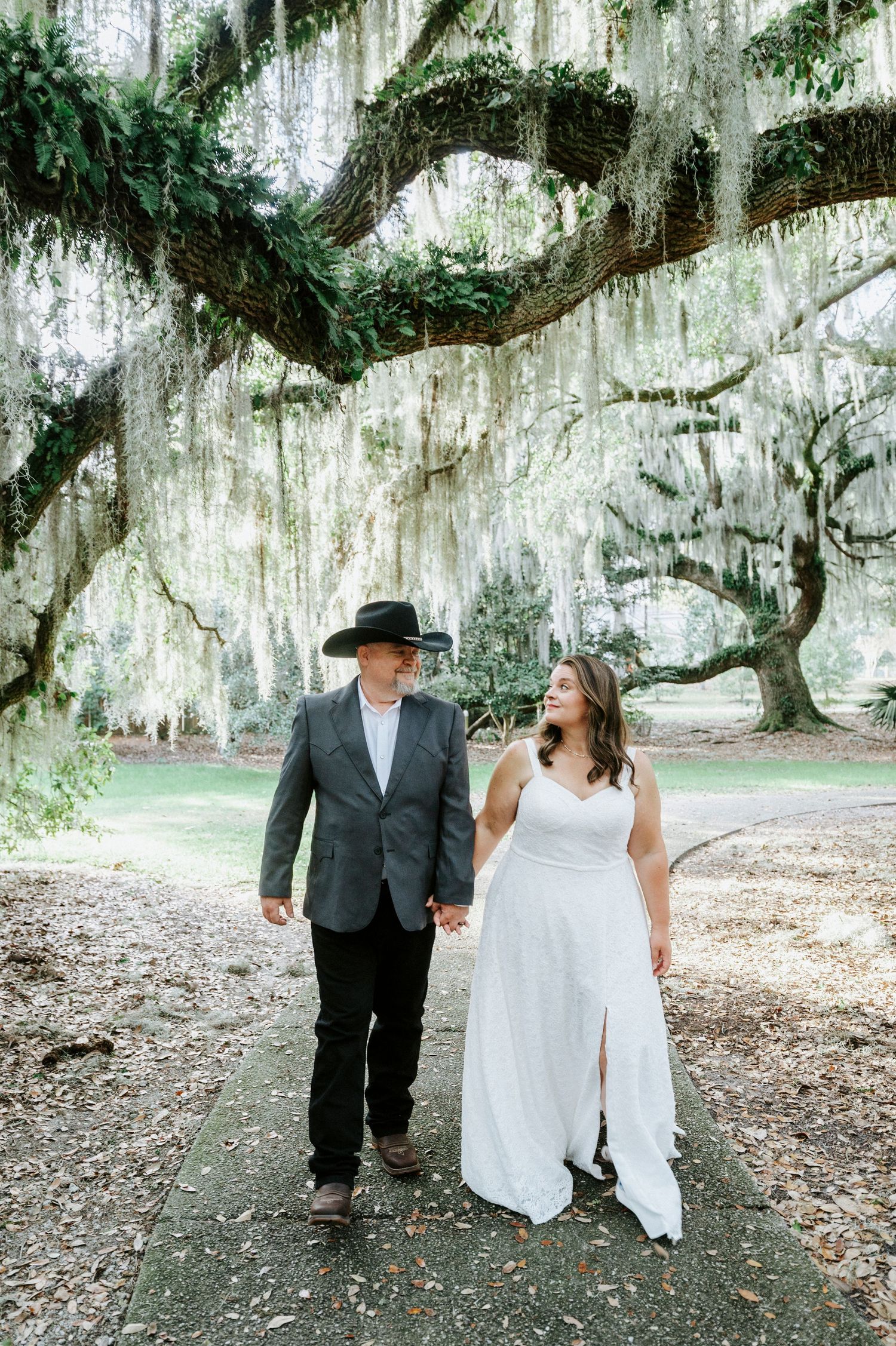 A couple walks together beneath majestic Spanish moss-draped oak trees in a romantic Southern garden setting.