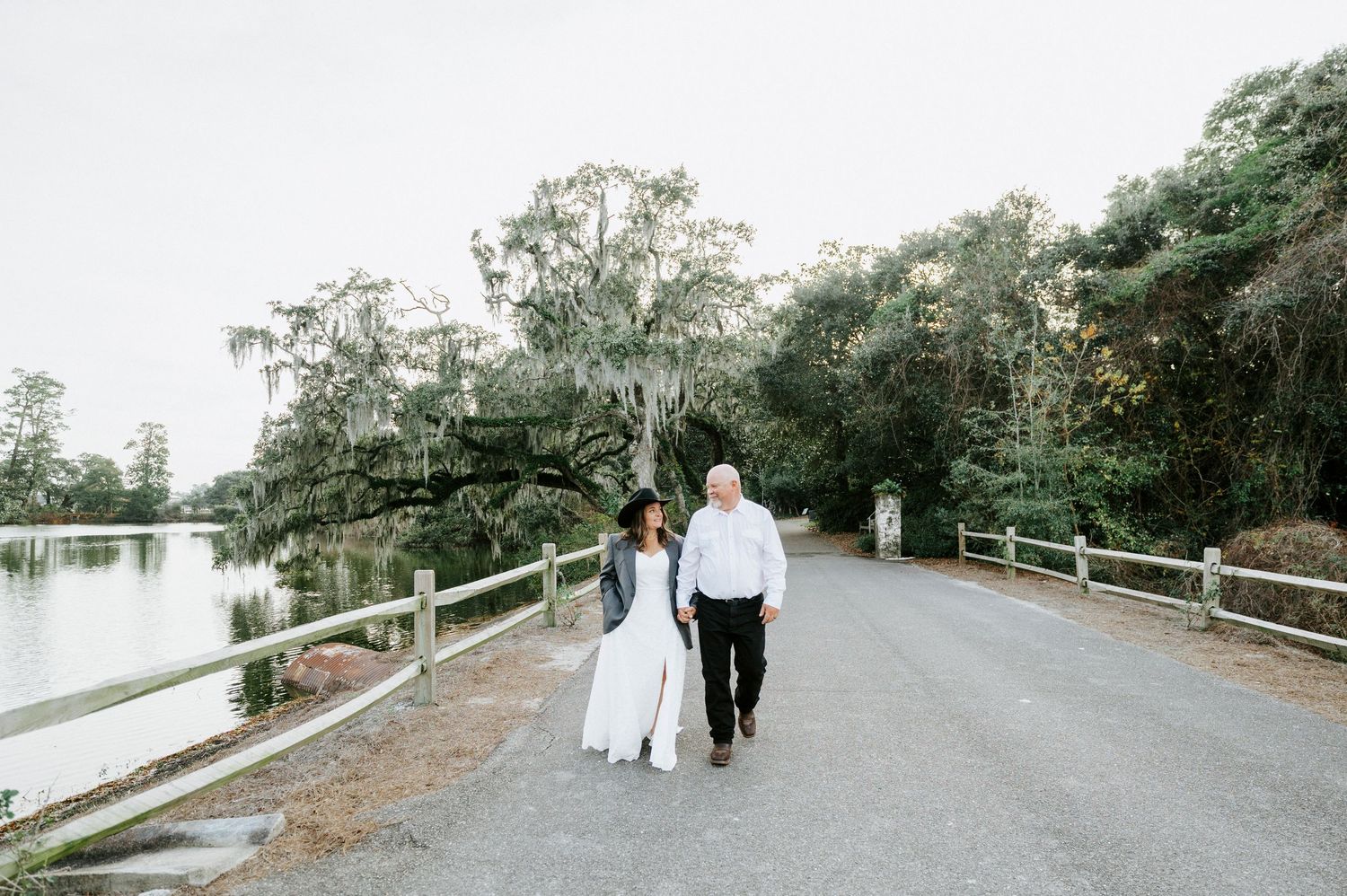 A couple walks hand in hand down a tree-lined path beside a tranquil lake.