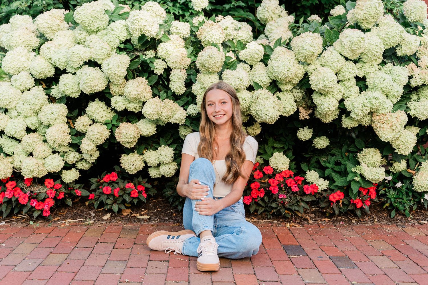 Senior photo against a backdrop of white hydrangea bushes and red flowers along a brick pathway in Grand Rapids, Ohio