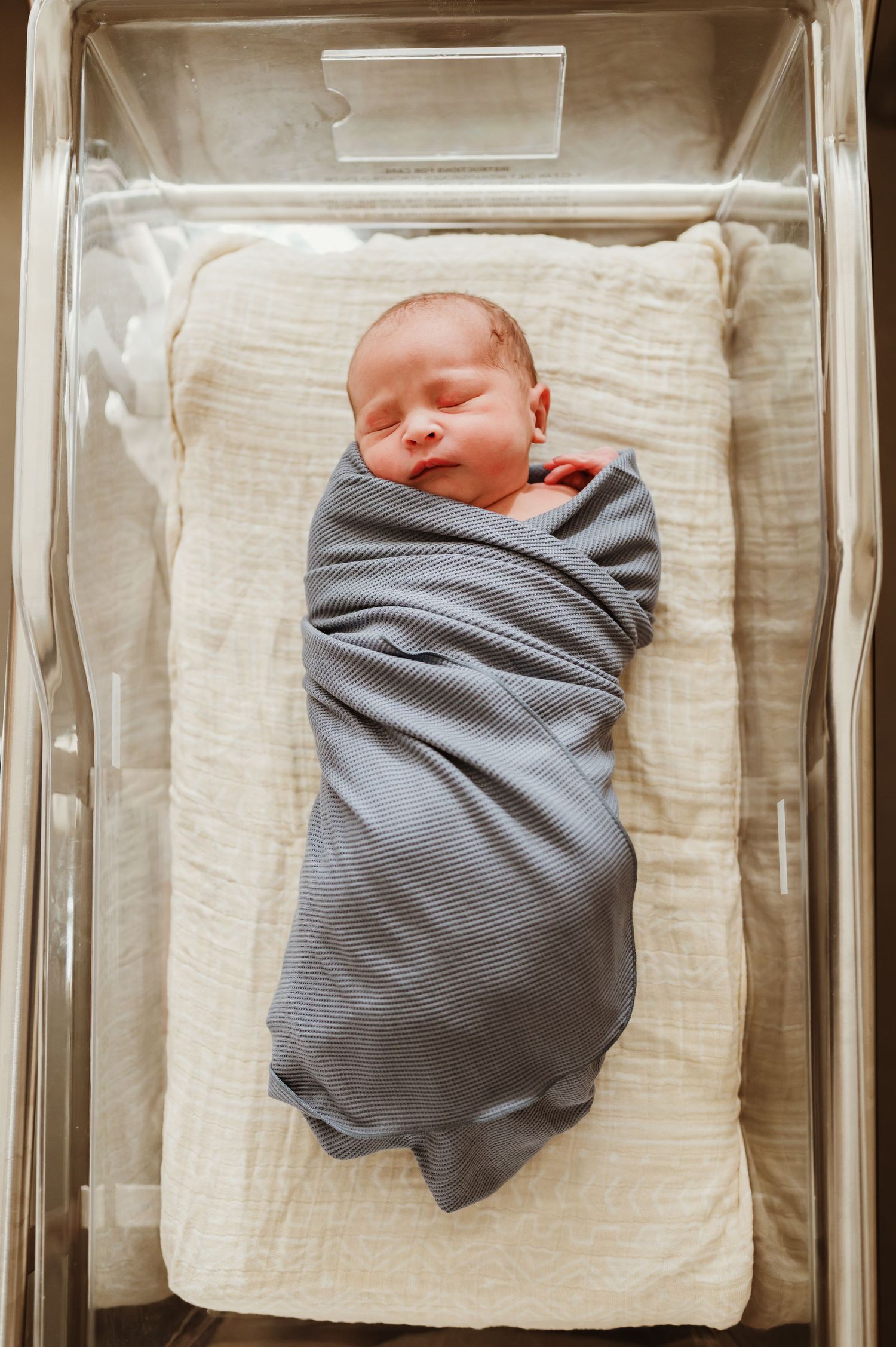 A newborn baby swaddled in gray blankets rest in hospital bassinets side by side.