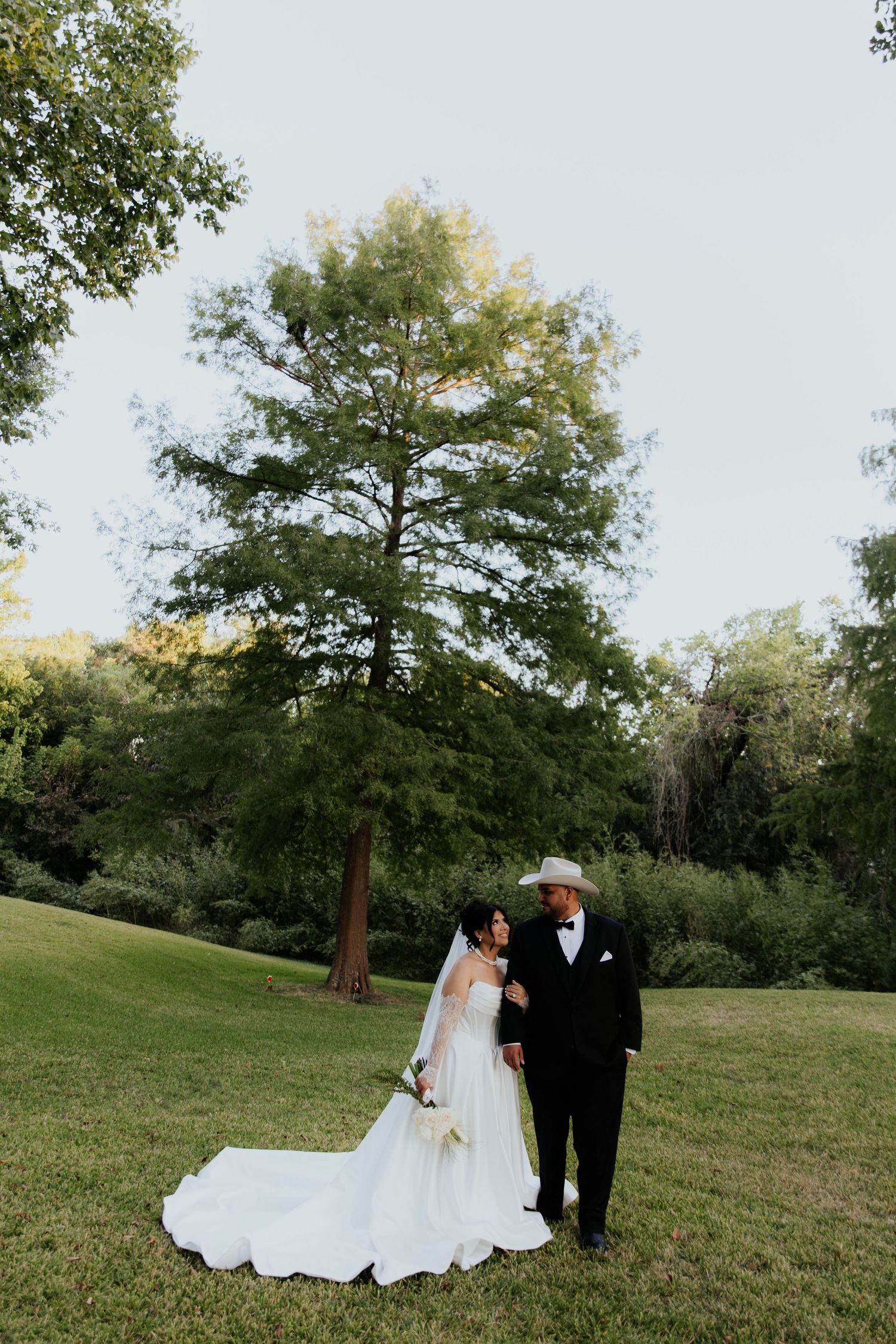 Elegant wedding couple poses on green lawn with trees in background during sunset.