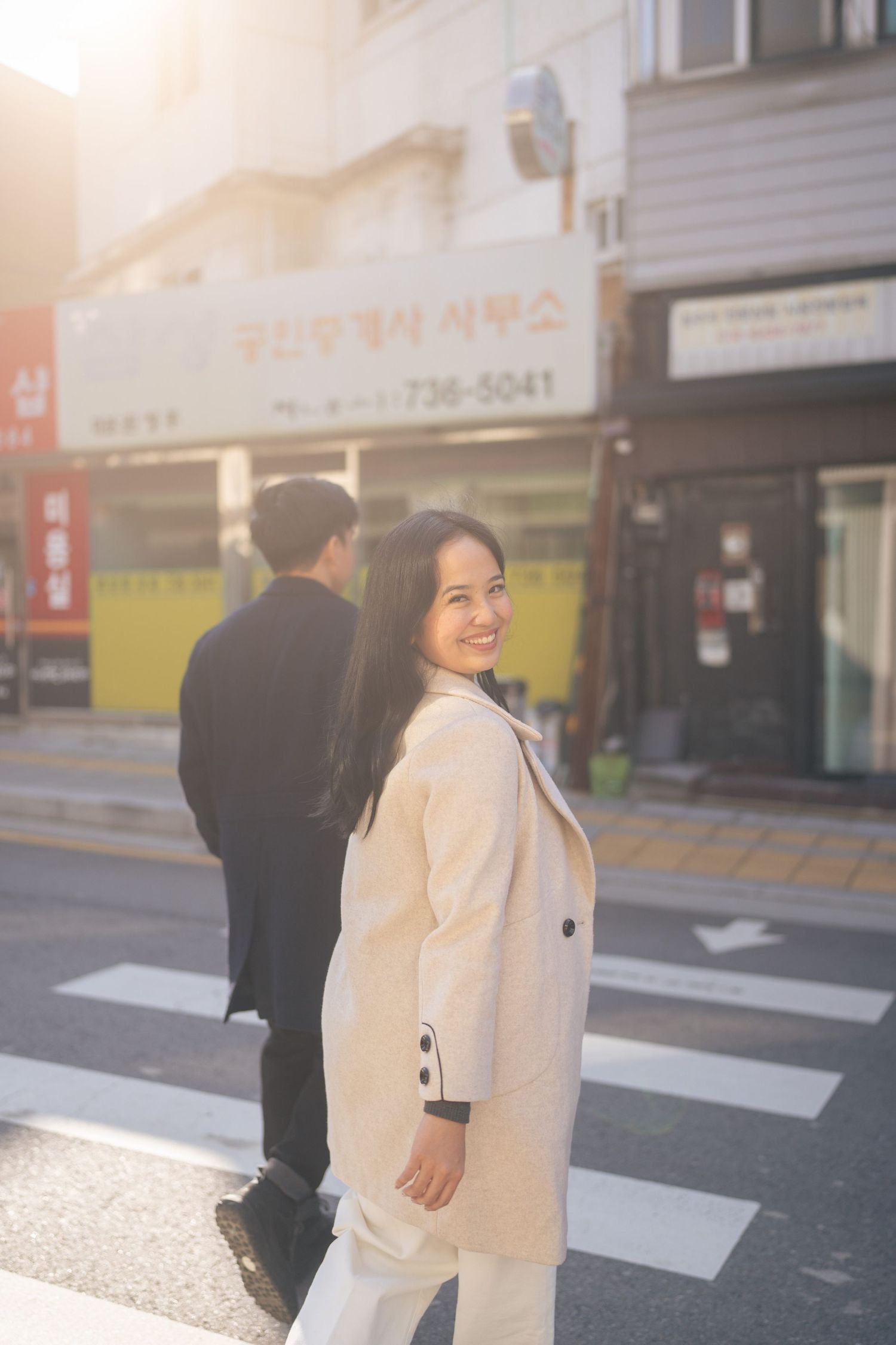 A person in a beige coat walks across a crosswalk on a city street during golden hour lighting.