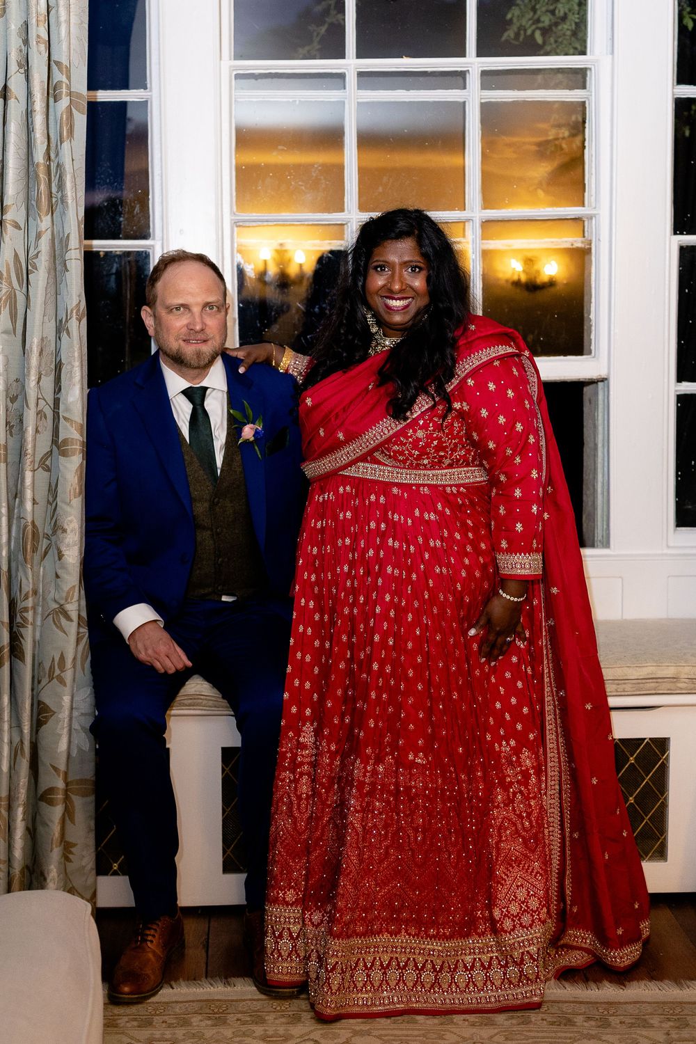 A couple poses for photos at a wedding celebration, with the bride wearing a stunning red and gold traditional Indian sari.
