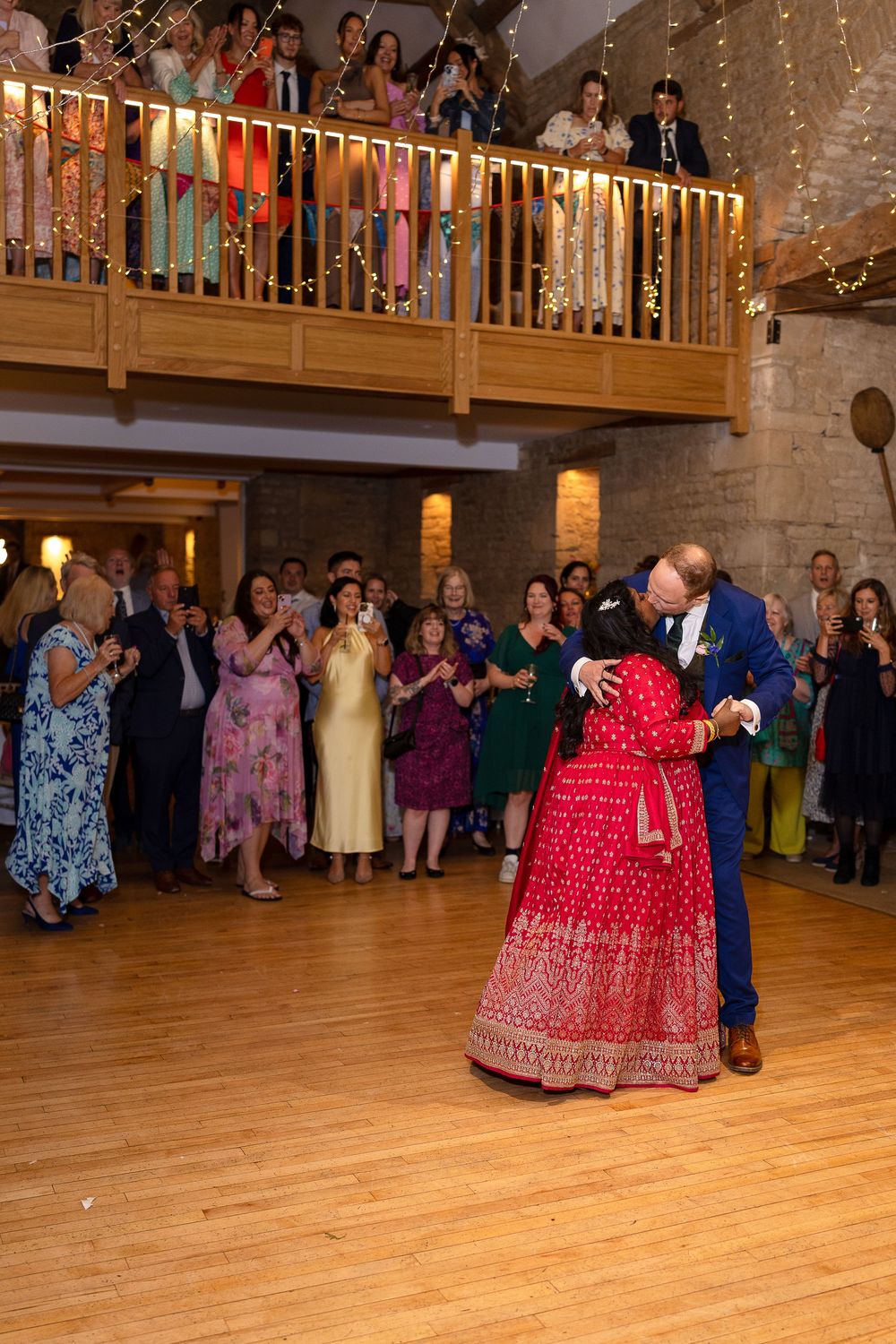 Wedding guests watch from a wooden balcony as a couple shares their first dance on a hardwood floor below.