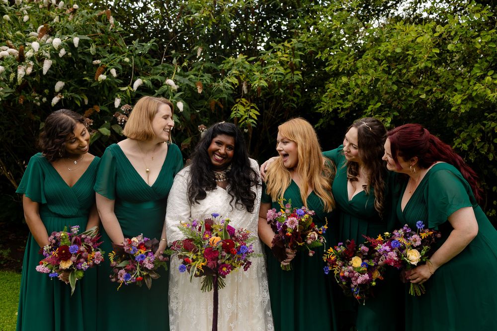 A wedding party in emerald green dresses holds vibrant wildflower bouquets while sharing a joyful moment in a garden setting.