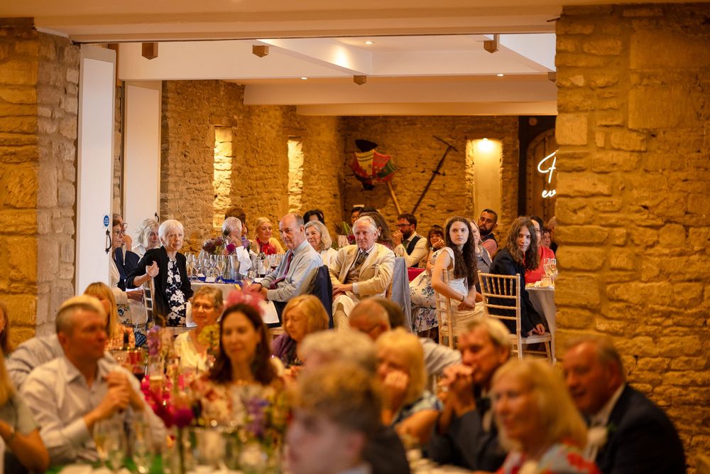 Wedding guests applaud during a reception in a rustic stone-walled venue with warm lighting.
