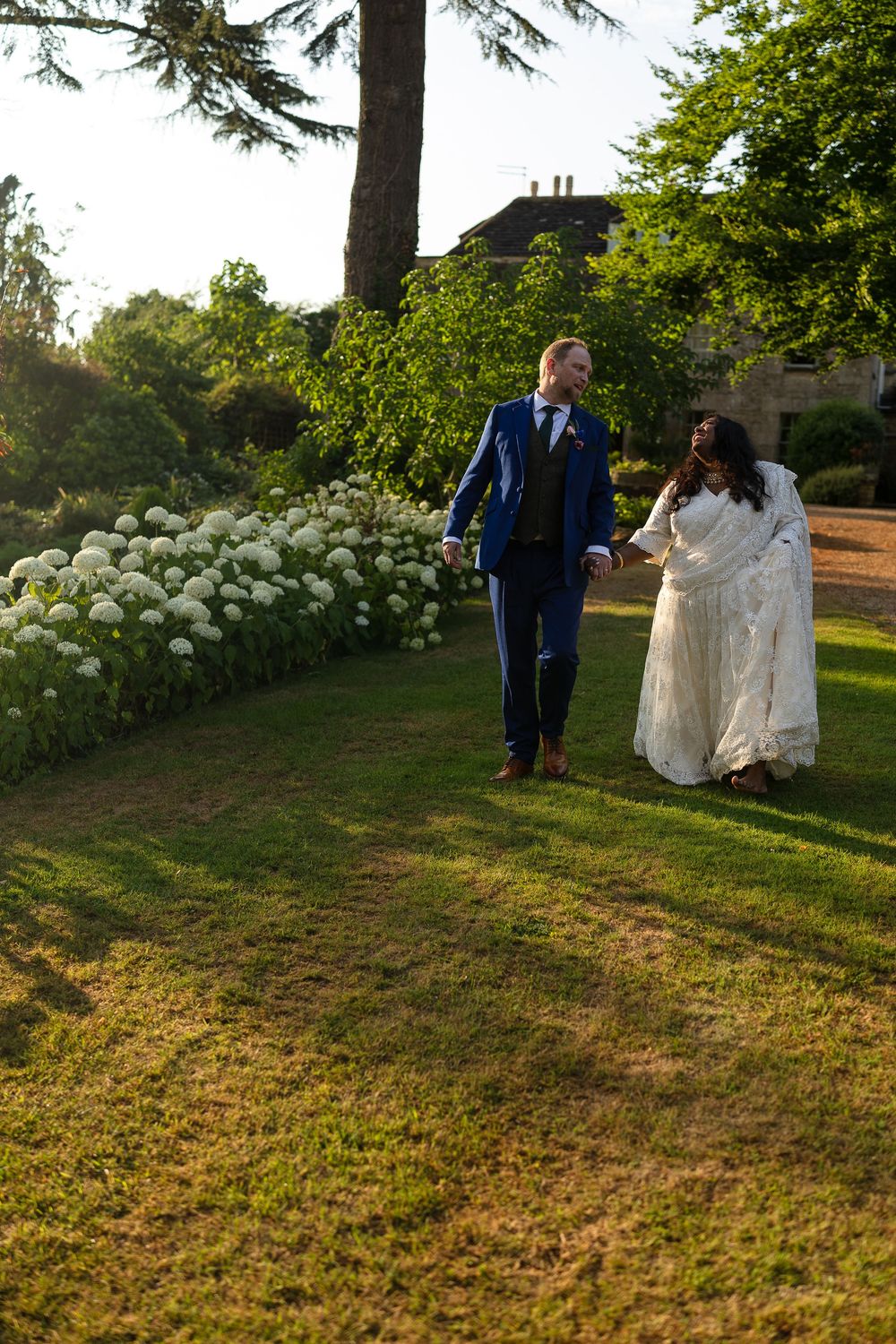 A couple walks along a garden path lined with blooming white hydrangea bushes on a sunny summer evening.