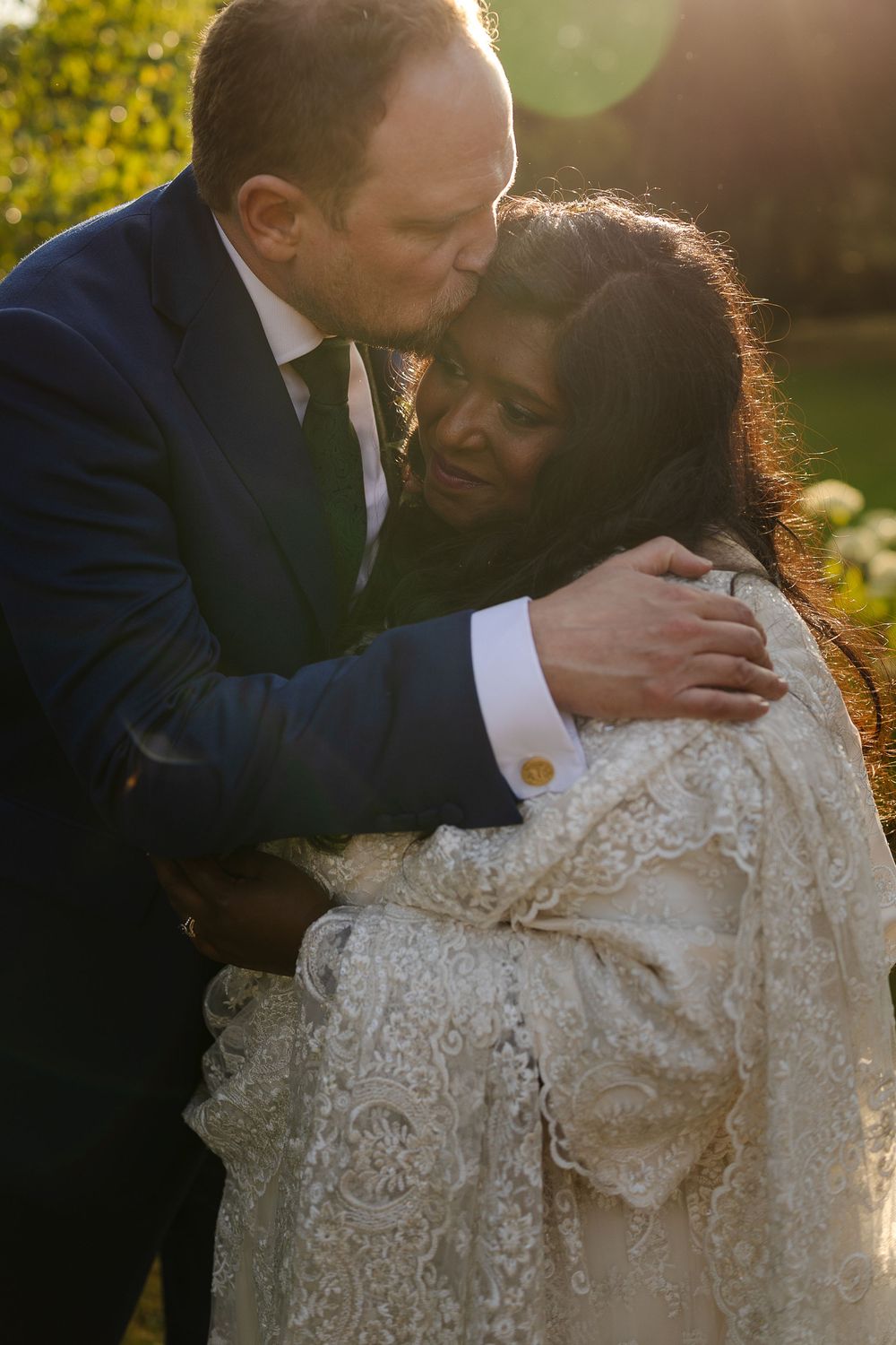 Couple embraces during golden hour outdoor wedding photoshoot with sunflare and lace details.