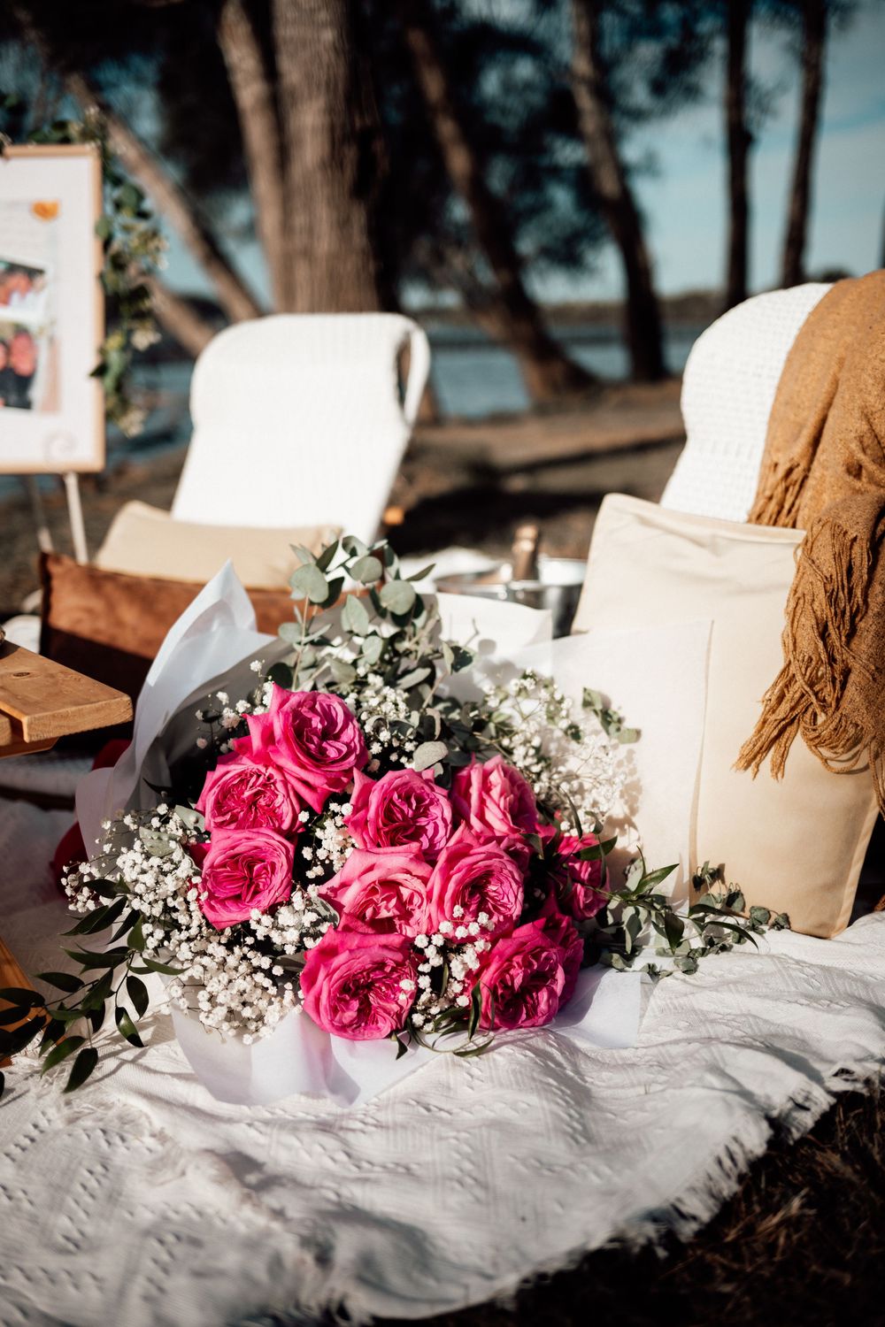 Pink roses and baby's breath flowers arranged on white linens with rustic outdoor seating in the background.