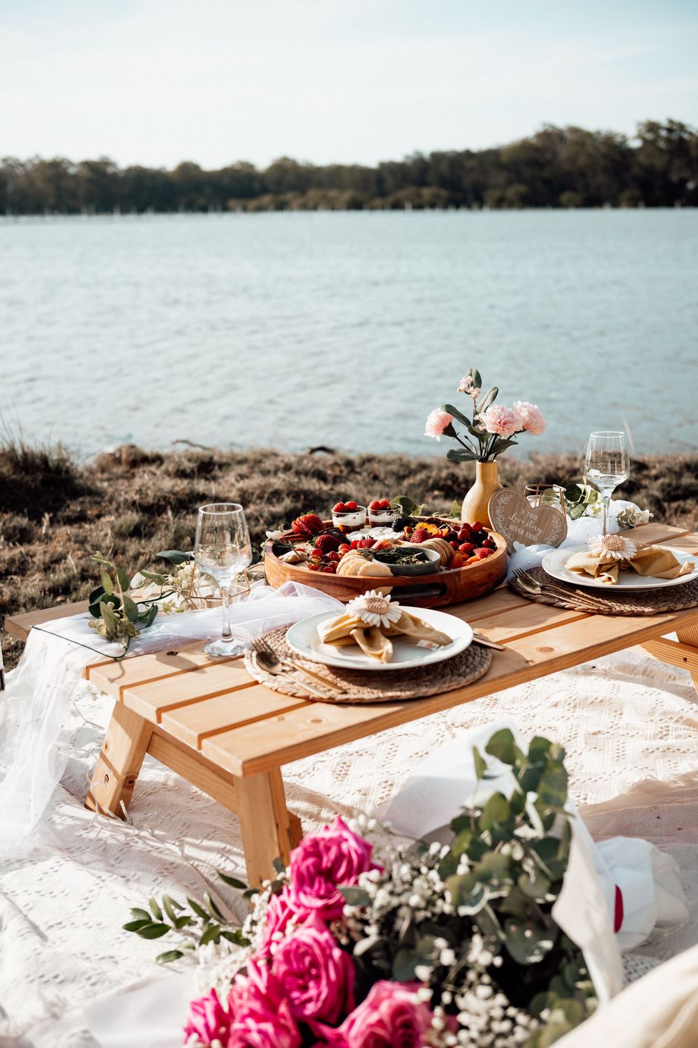 Romantic lakeside picnic setup with wooden tray, wine glasses, and pink roses on white blanket.
