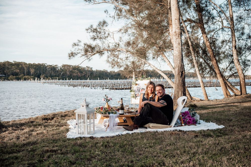 A romantic picnic setup with blankets and pillows under tall pine trees beside a tranquil lake at sunset.