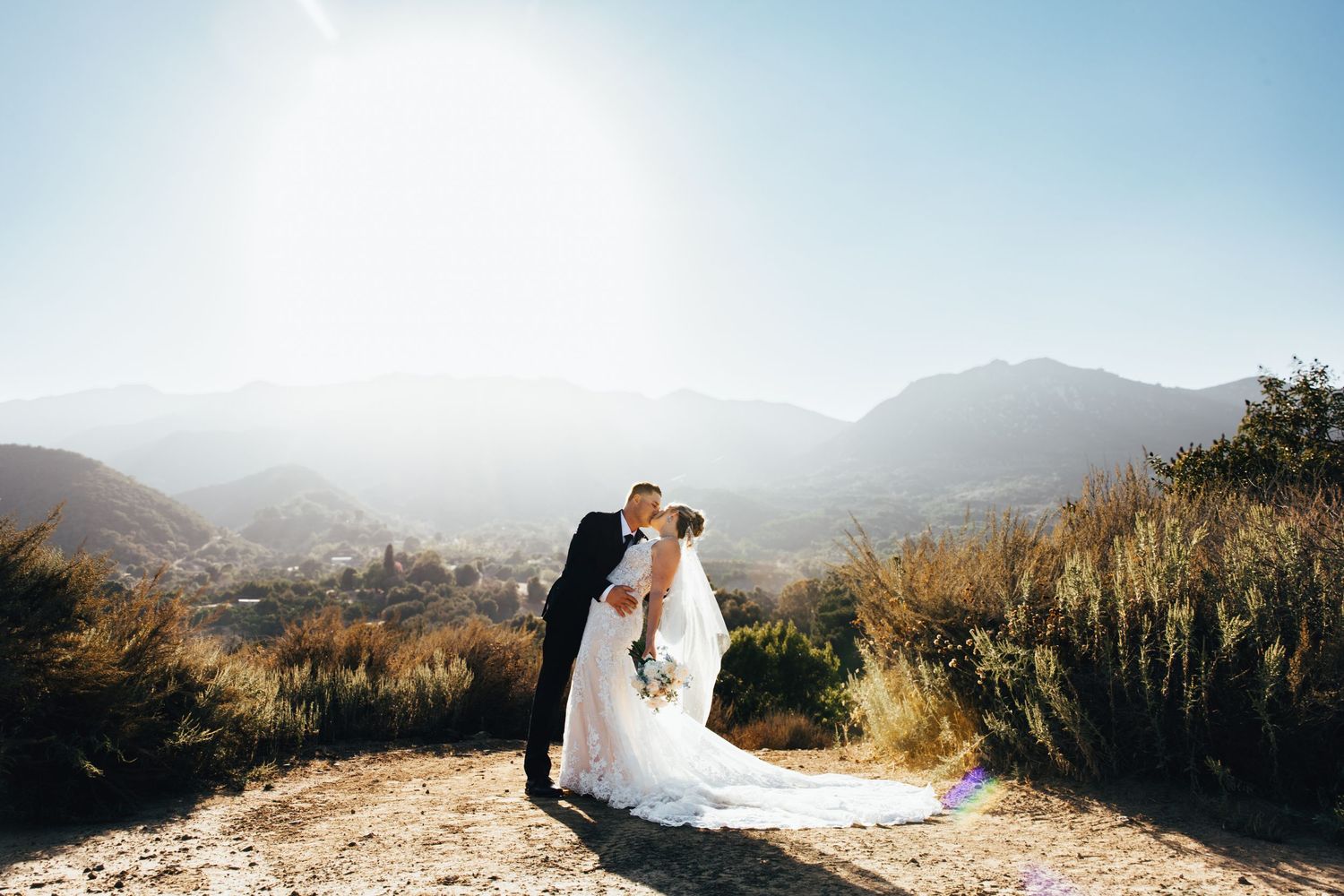 A couple shares a romantic moment at sunset on a mountain overlook with a scenic valley vista in the background.